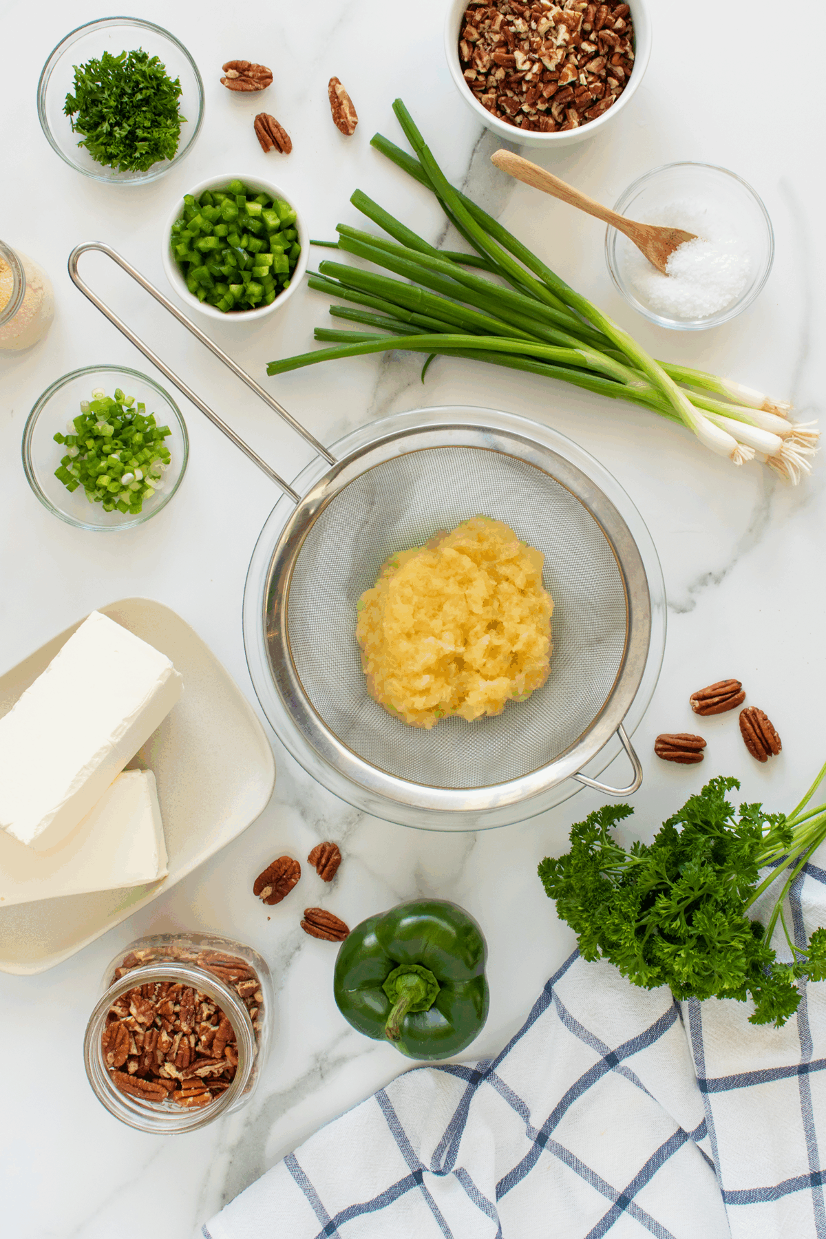 Ingredients including cream cheese, crushed pineapple in a strainer, green onions, chopped parsley, bell pepper, pecans, and salt are arranged on a white surface with a kitchen towel.