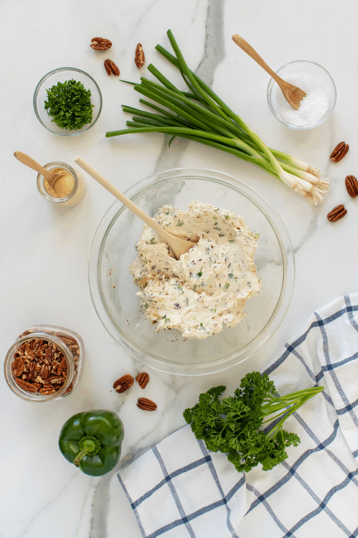 A glass bowl of herbed cream cheese mixture with a wooden spoon, surrounded by chopped pecans, green onions, parsley, green bell pepper, a jar of pecans, garlic powder, salt, and a blue-striped kitchen towel.