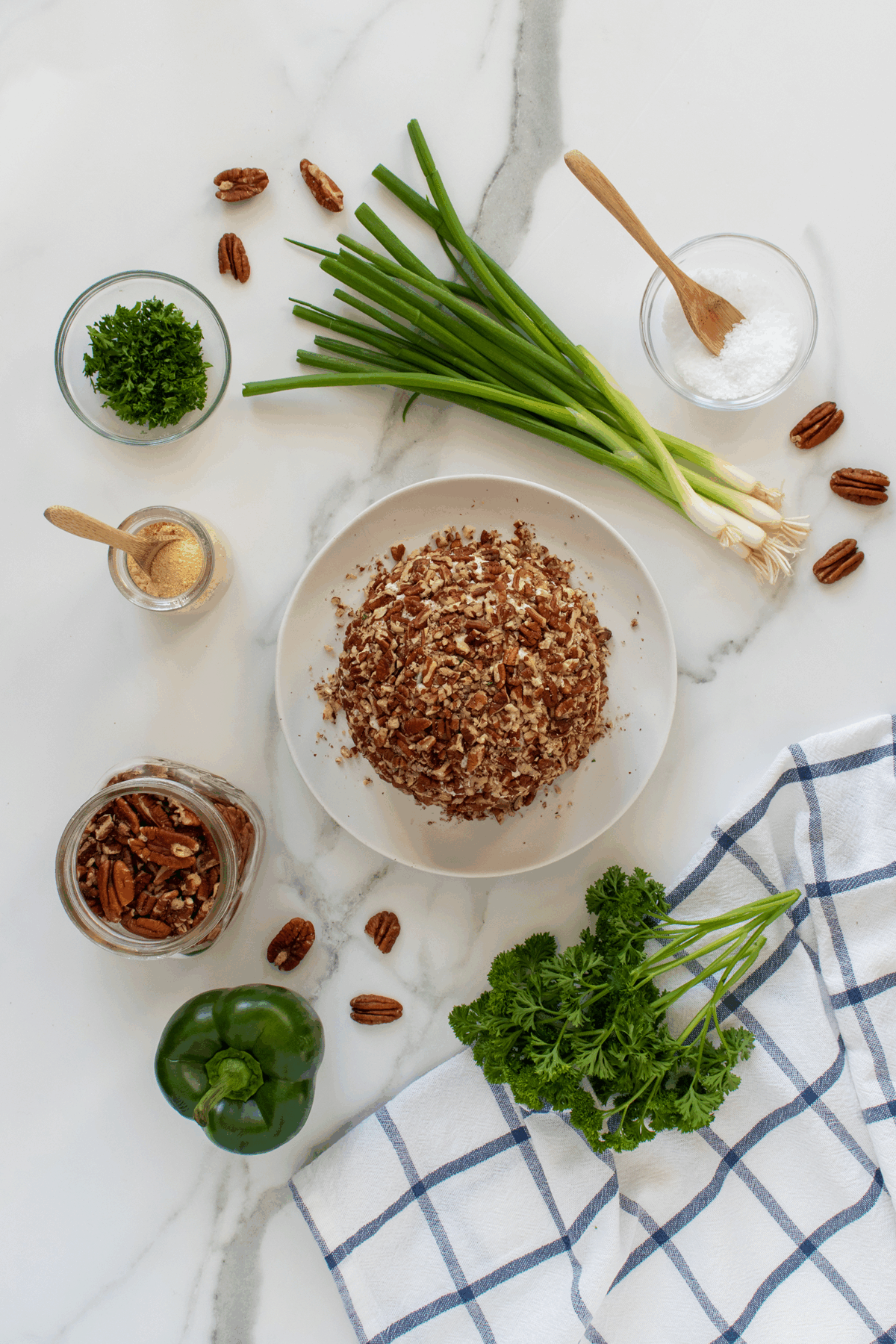 A cheese ball coated with chopped pecans sits on a plate, surrounded by green onions, fresh parsley, a green bell pepper, pecans, a jar of pecans, salt, ground seasoning, and a checkered kitchen towel.