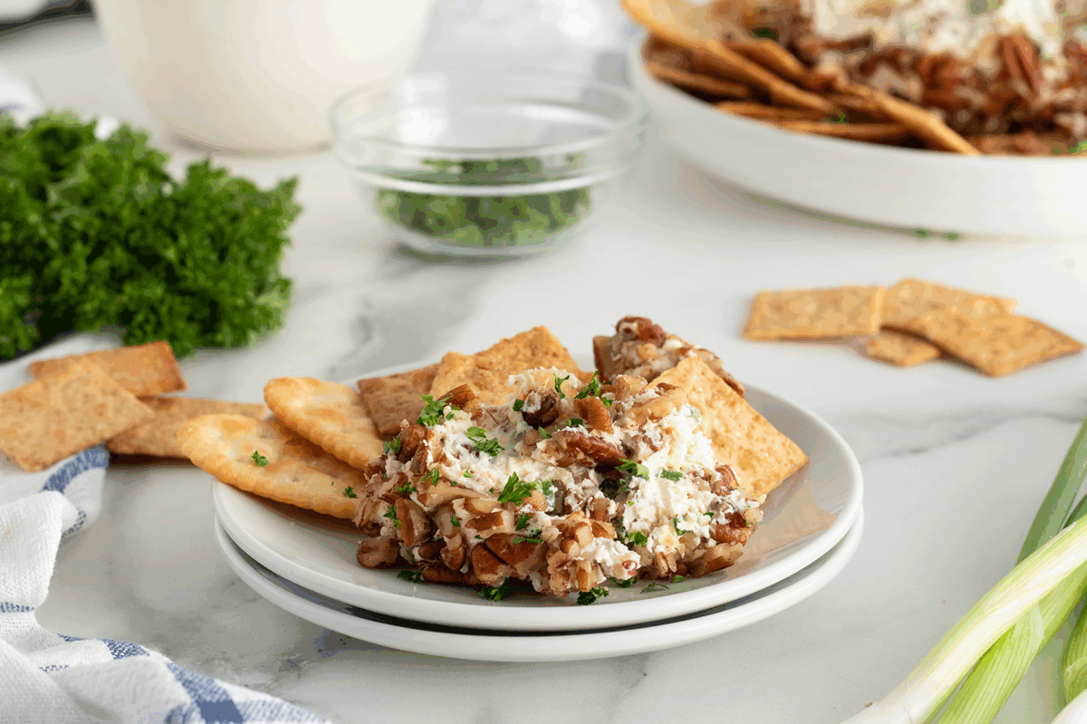 A plate of crackers with a serving of cheese spread topped with chopped pecans and herbs. Fresh parsley, green onions, and more crackers are in the background on a white marble surface.