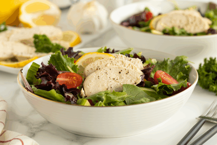 A bowl of mixed green salad topped with sliced cooked chicken breast, cherry tomatoes, and a lemon wedge. Another bowl of salad and slices of bread are in the background.