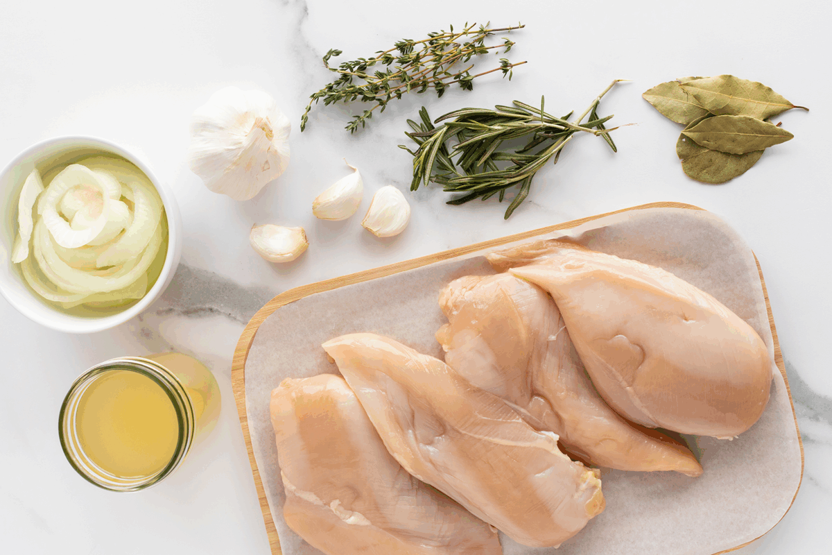Raw chicken breasts on a wooden board beside a bowl of sliced onions, garlic bulbs and cloves, fresh thyme, rosemary, bay leaves, and a jar of broth on a white marble surface.
