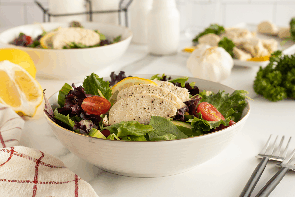 A bowl of mixed green salad topped with sliced grilled chicken breast, cherry tomatoes, and a lemon wedge sits on a white table beside a fork, a lemon, garlic, and a checkered napkin.