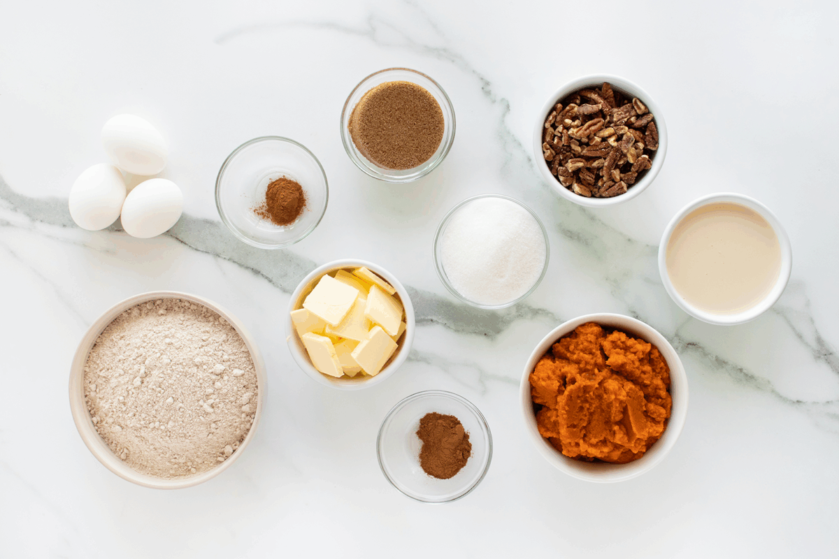 A top-down view of baking ingredients on a white marble surface, including eggs, flour, butter, sugar, brown sugar, spices, pecans, milk, and a bowl of pumpkin puree.