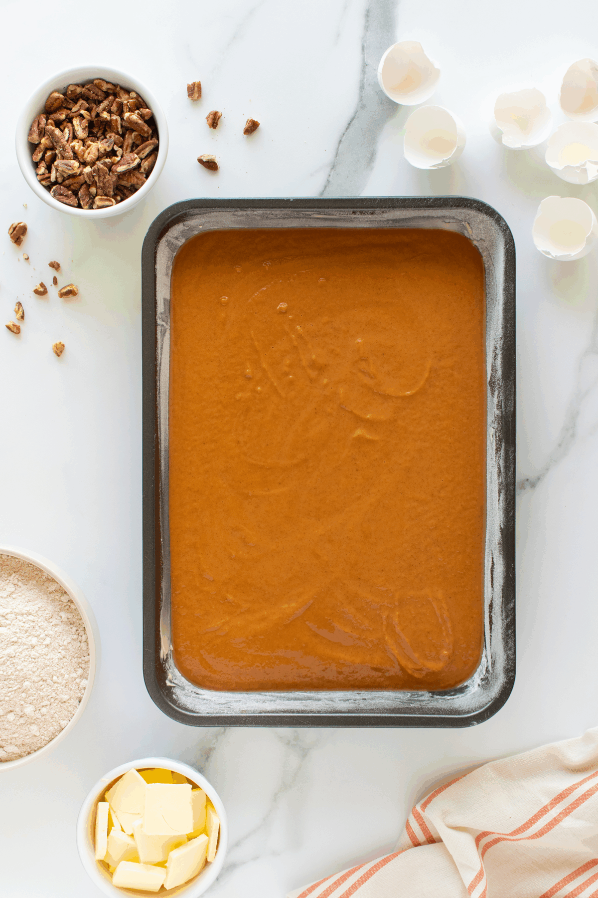 A baking tray filled with pumpkin batter sits on a marble surface, surrounded by bowls of chopped pecans, flour, and butter, with eggshells scattered nearby and a striped towel in the corner.