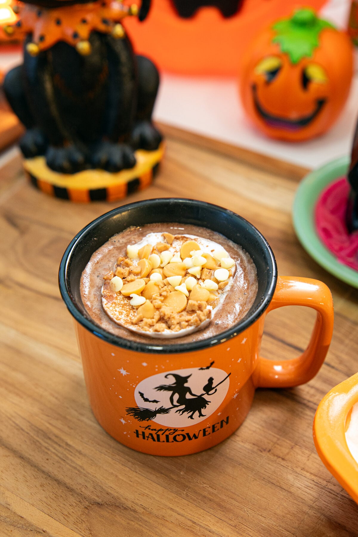 An orange mug with a Halloween witch design filled with hot chocolate topped with whipped cream and white chocolate chips, on a wooden surface. Halloween decorations, including a jack-o-lantern, are in the background.