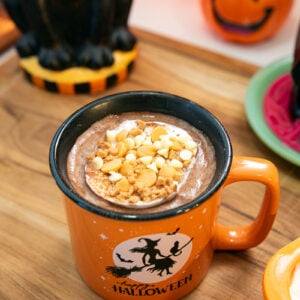 An orange mug with a Halloween witch design filled with hot chocolate topped with whipped cream and white chocolate chips, on a wooden surface. Halloween decorations, including a jack-o-lantern, are in the background.
