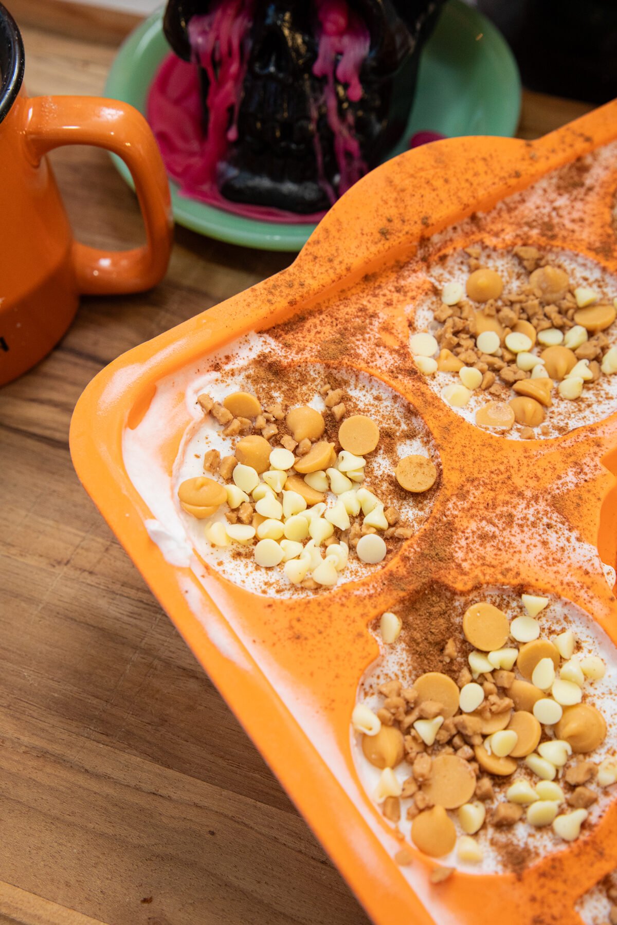 An orange tray holds desserts topped with white and butterscotch chips and cinnamon. An orange mug and a black skull candle with melted pink wax sit nearby on a wooden surface.