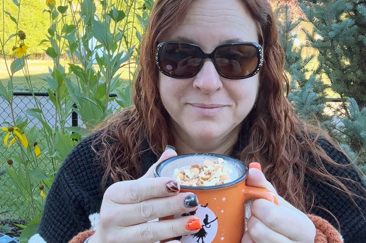 A woman with wavy red hair and sunglasses smiles while holding an orange mug with a witch design. She has Halloween-themed nail art and sits outdoors with green plants in the background.