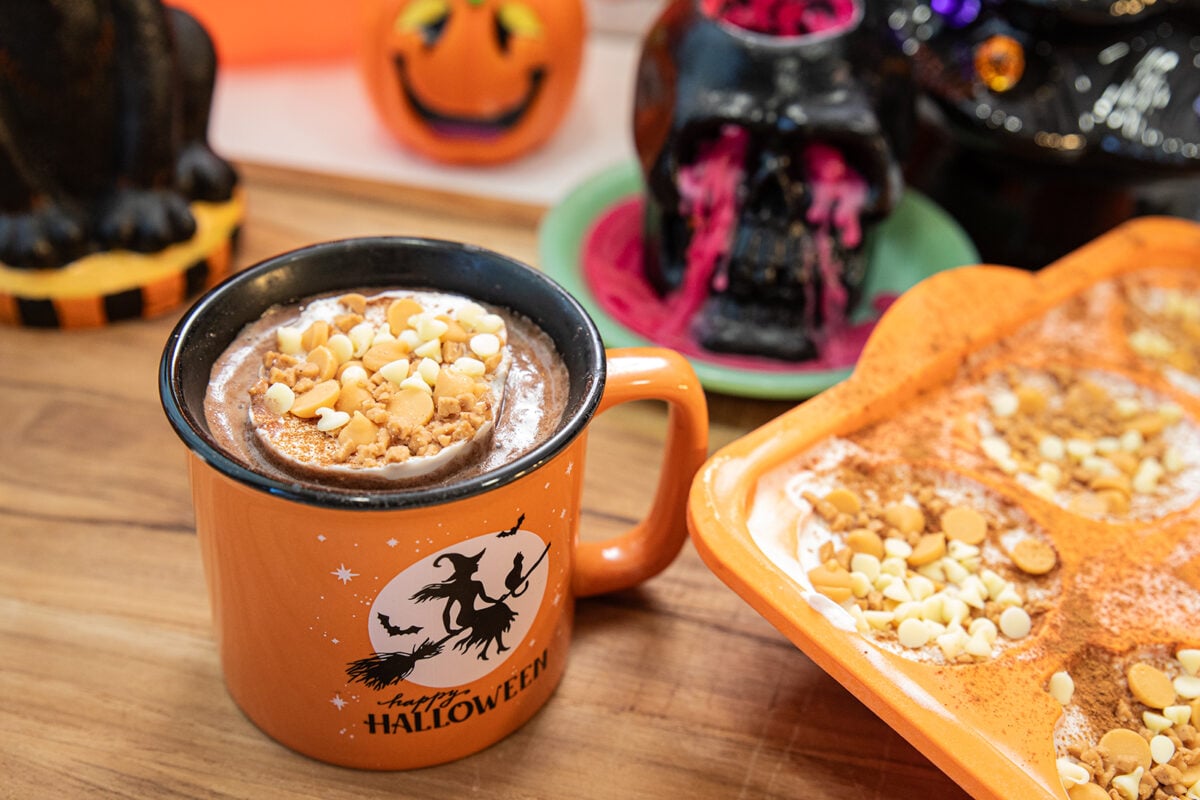 An orange mug with a witch and “Happy Halloween” filled with hot chocolate, topped with whipped cream, crumbled cookie, and white chocolate chips, sits on a wooden table with Halloween decorations in the background.