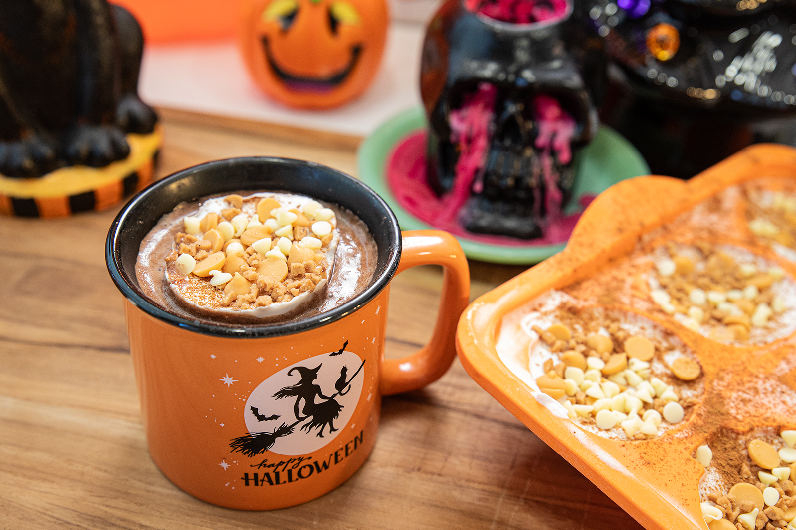 An orange mug with a witch and “Happy Halloween” filled with hot chocolate, topped with whipped cream, crumbled cookie, and white chocolate chips, sits on a wooden table with Halloween decorations in the background.