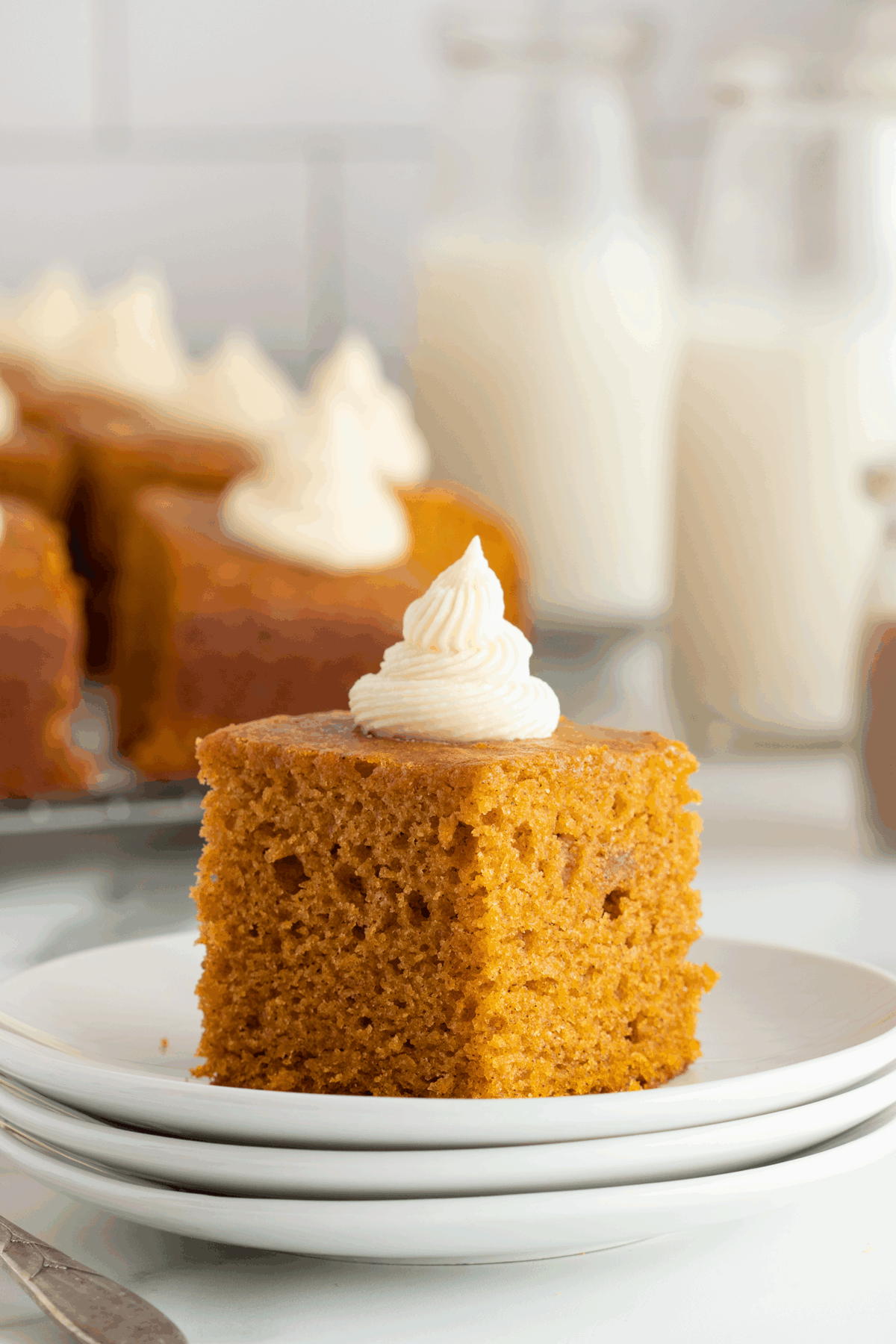 A slice of moist pumpkin cake with swirled cream cheese frosting sits on a stack of white plates. More cake pieces and bottles of milk are blurred in the background.