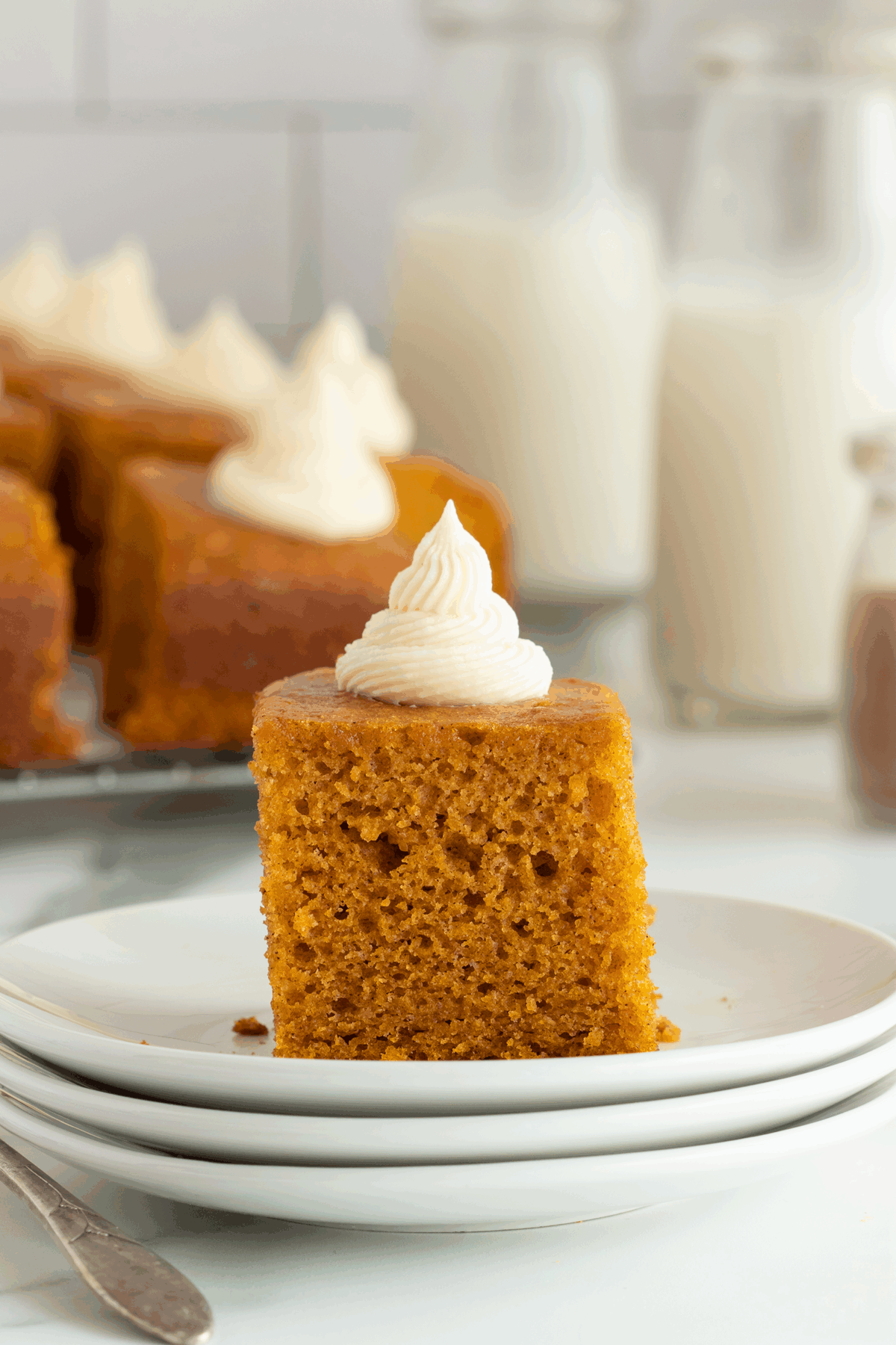 A slice of moist pumpkin cake topped with a swirl of white frosting sits on a stack of white plates, with more cake pieces and bottles of milk in the background.