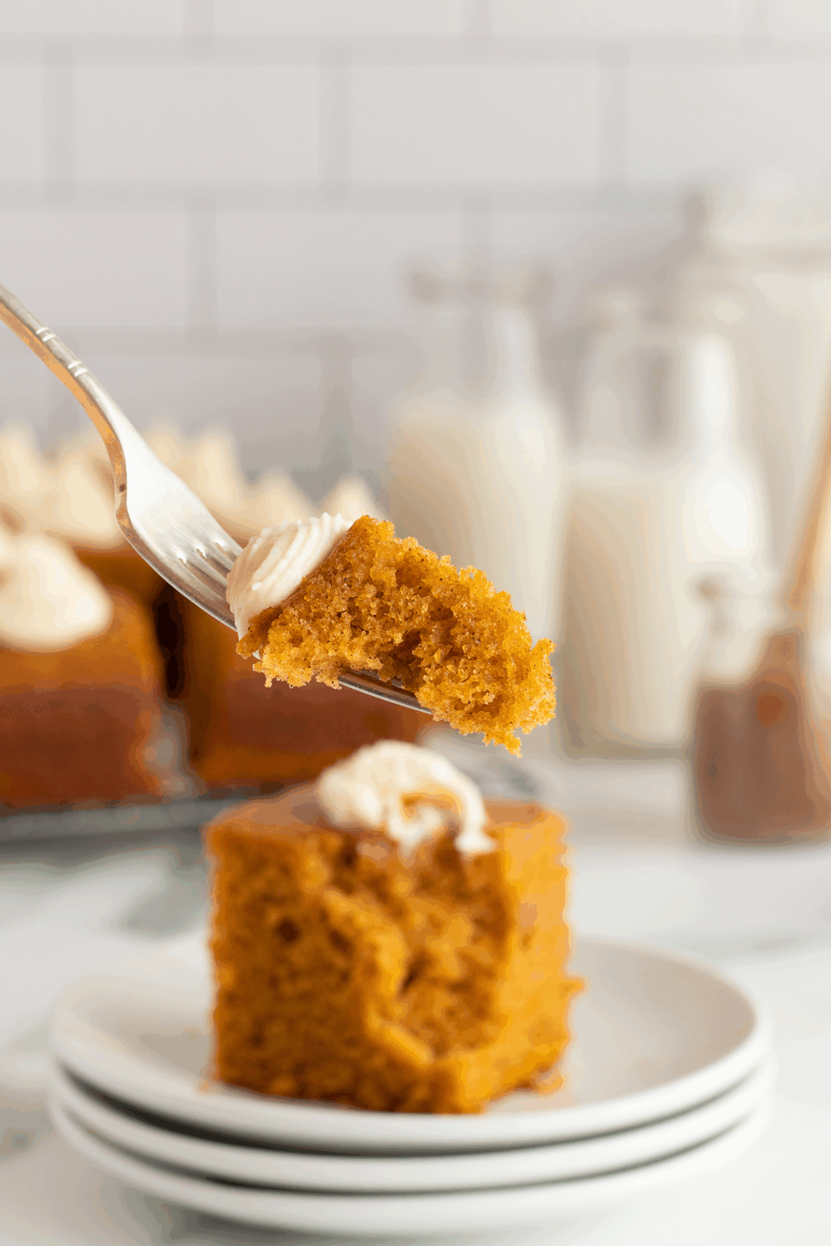 A fork holds a bite of moist, orange-brown cake with frosting, above a square slice of the same cake sitting on stacked white plates. Blurry bottles of milk and more cake appear in the background.