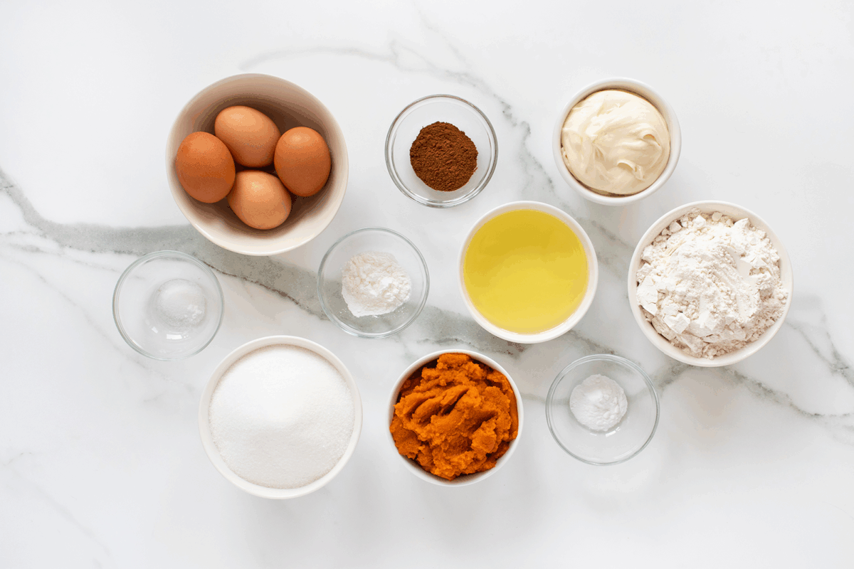 A top-down view of baking ingredients in bowls on a white marble surface, including eggs, cinnamon, cream cheese, vegetable oil, flour, sugar, pumpkin puree, baking soda, baking powder, and two empty bowls.