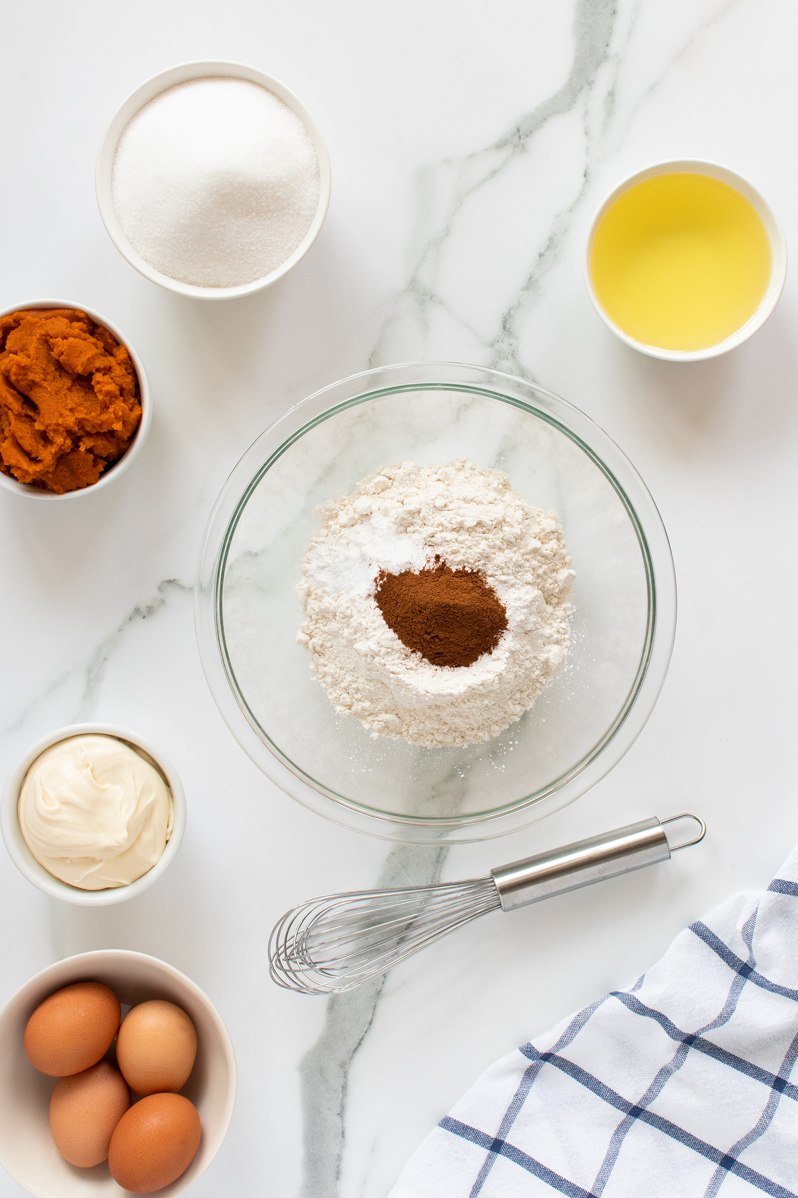 A bowl of flour and cinnamon is surrounded by bowls of sugar, oil, pumpkin puree, eggs, and cream cheese on a marble countertop. A metal whisk and a blue-and-white towel are nearby.