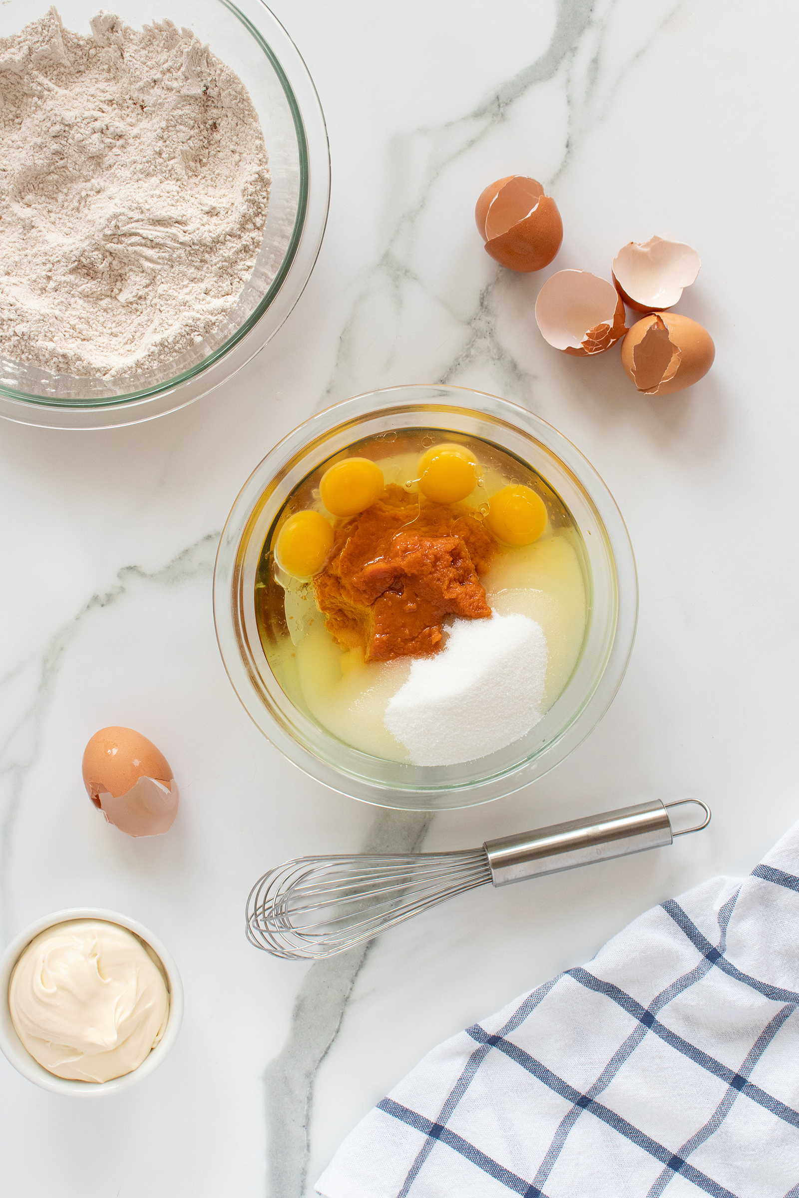 A mixing bowl with eggs, pumpkin puree, sugar, and oil, surrounded by cracked eggshells, flour in another bowl, a small bowl of mayonnaise, a whisk, and a checkered kitchen towel on a marble surface.