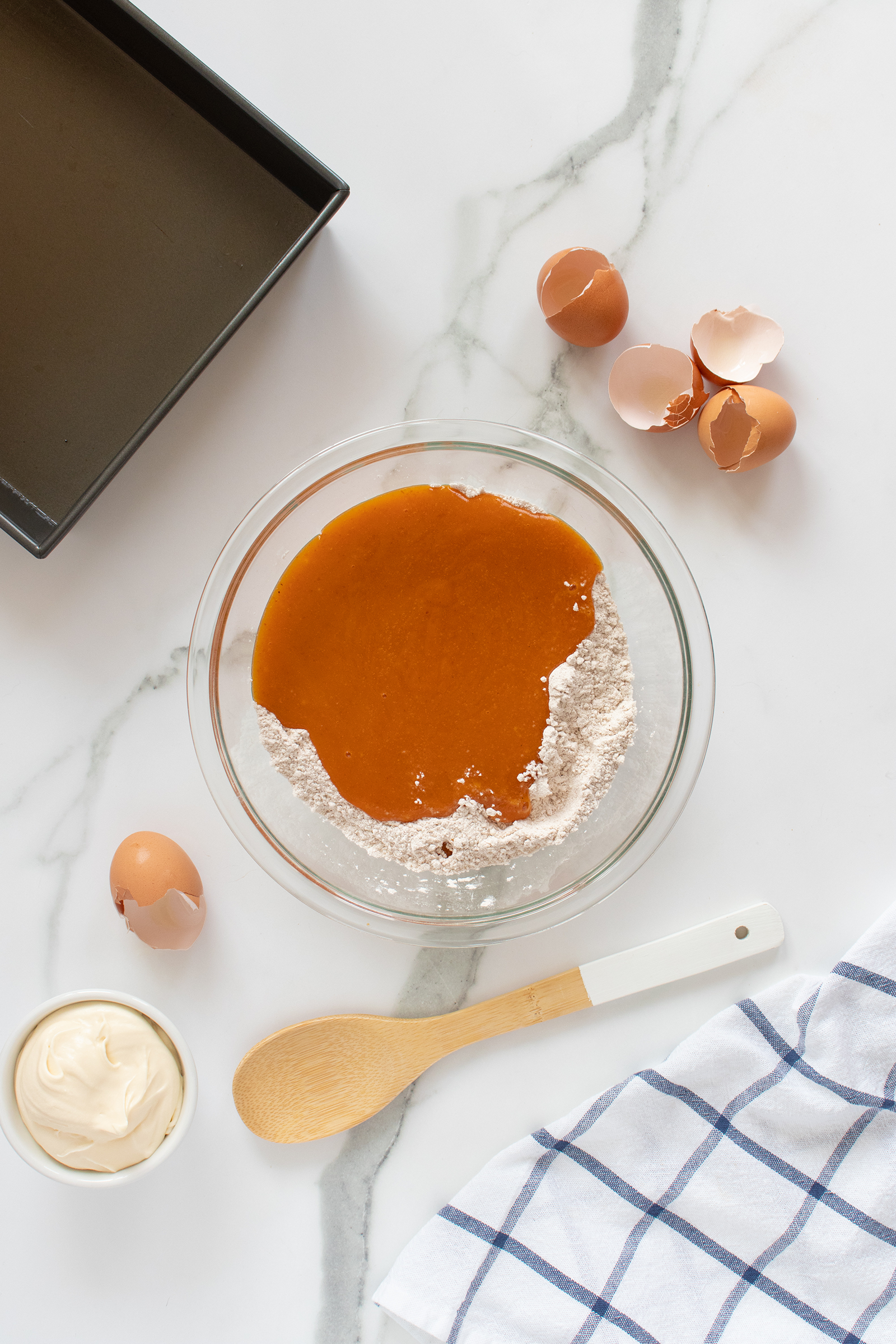 A glass bowl with flour and pumpkin puree sits on a marble surface next to cracked eggshells, a square baking pan, a small dish of cream cheese, a wooden spoon, and a blue and white checkered towel.