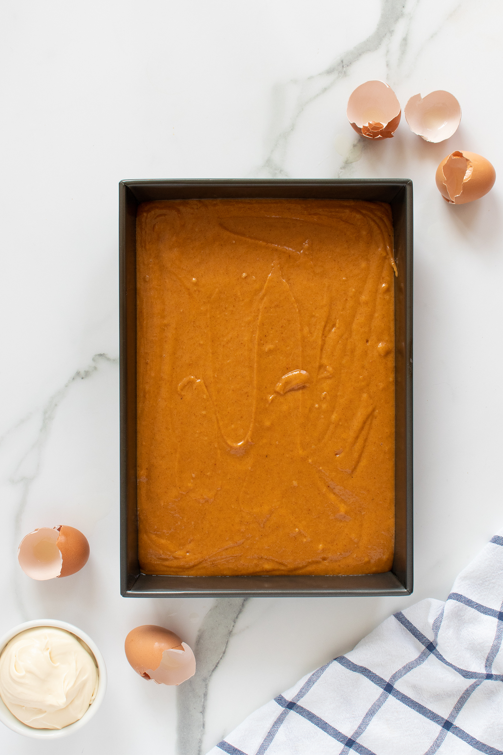 A rectangular baking pan filled with brown cake batter sits on a marble surface. Broken eggshells, a small bowl of cream, and part of a blue-checkered cloth are nearby.
