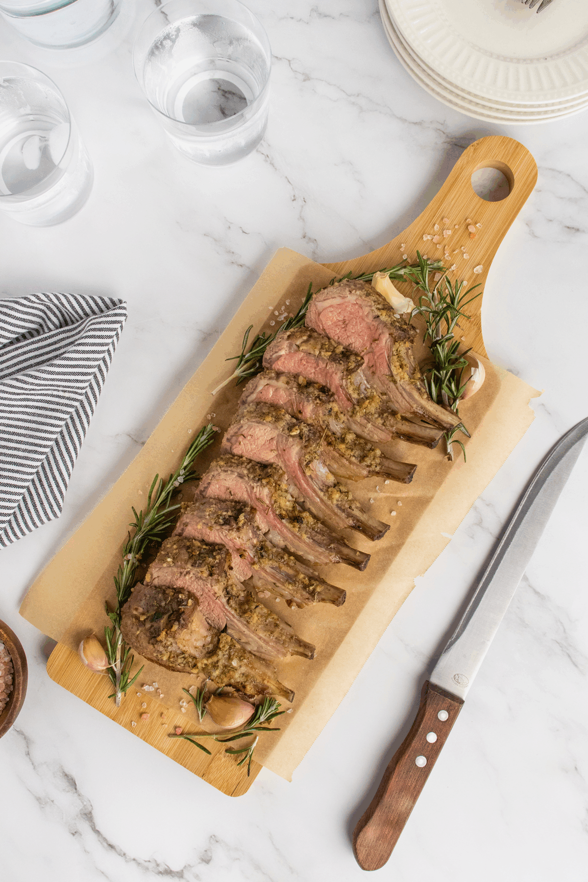 A sliced rack of herb-crusted lamb is arranged on a wooden cutting board with parchment paper, garnished with fresh rosemary and garlic. A knife, glass of water, plates, and a striped napkin are nearby on a marble countertop.