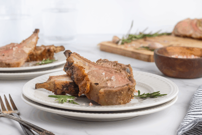 Two slices of cooked rack of lamb garnished with rosemary are served on a white plate, with another plate, a wooden bowl, and a cutting board with more lamb and rosemary in the background.