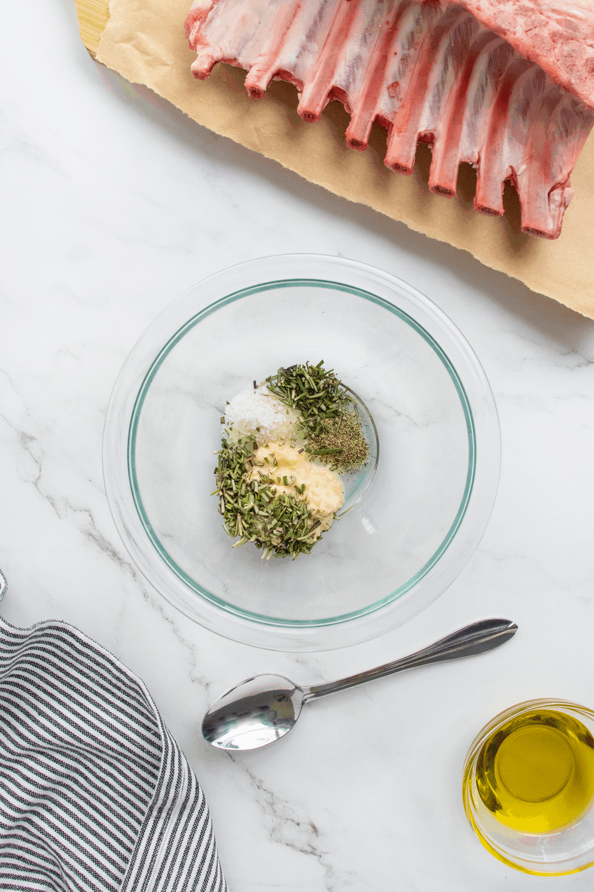 A glass bowl with chopped herbs, minced garlic, salt, pepper, and mustard sits on a marble surface near a rack of raw lamb, a spoon, a striped towel, and a small glass container of olive oil.
