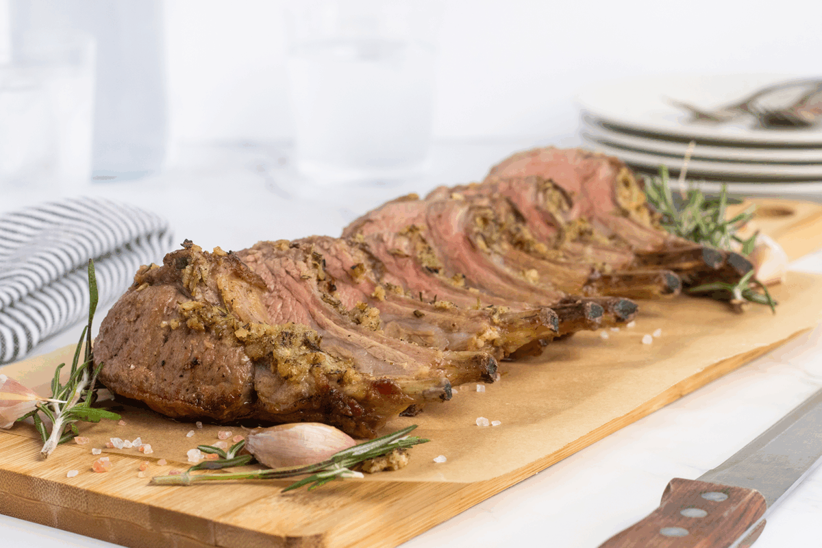 A sliced rack of lamb with a herb crust is served on a wooden board, garnished with rosemary and garlic, with a striped napkin and a stack of plates and cutlery in the background.