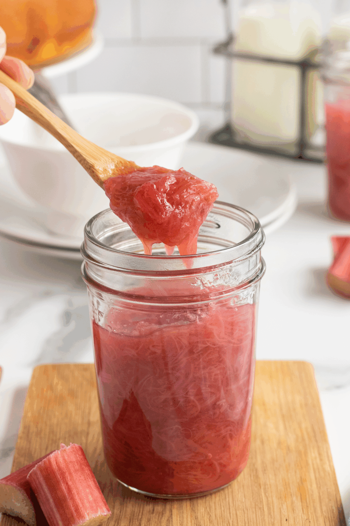 A wooden spoon lifting thick, pink rhubarb compote from a glass jar on a wooden board, with fresh rhubarb pieces and kitchen items in the background.