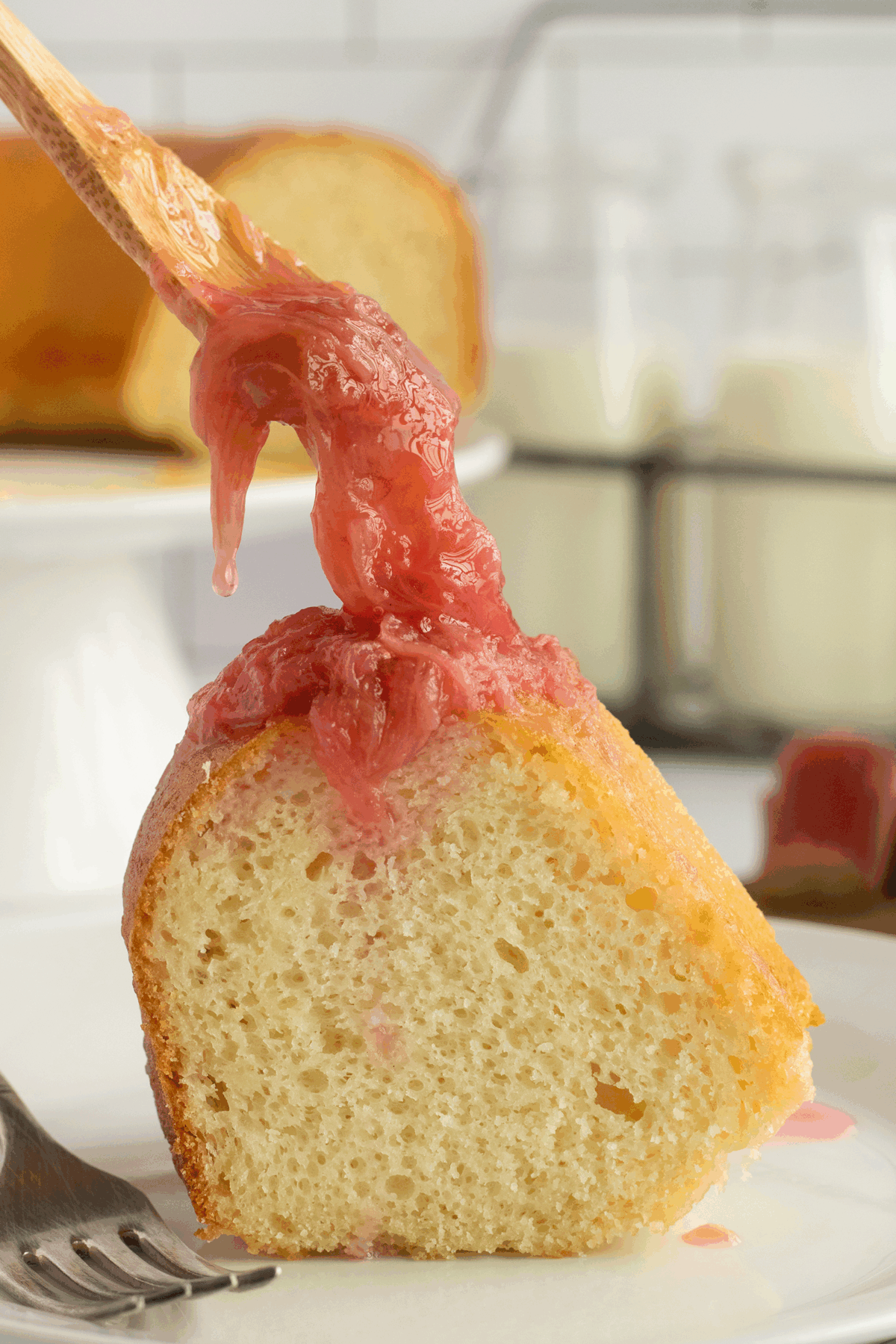A slice of bundt cake on a plate with pink rhubarb sauce being drizzled on top, a fork beside it, and bottles of milk and more cake in the background.