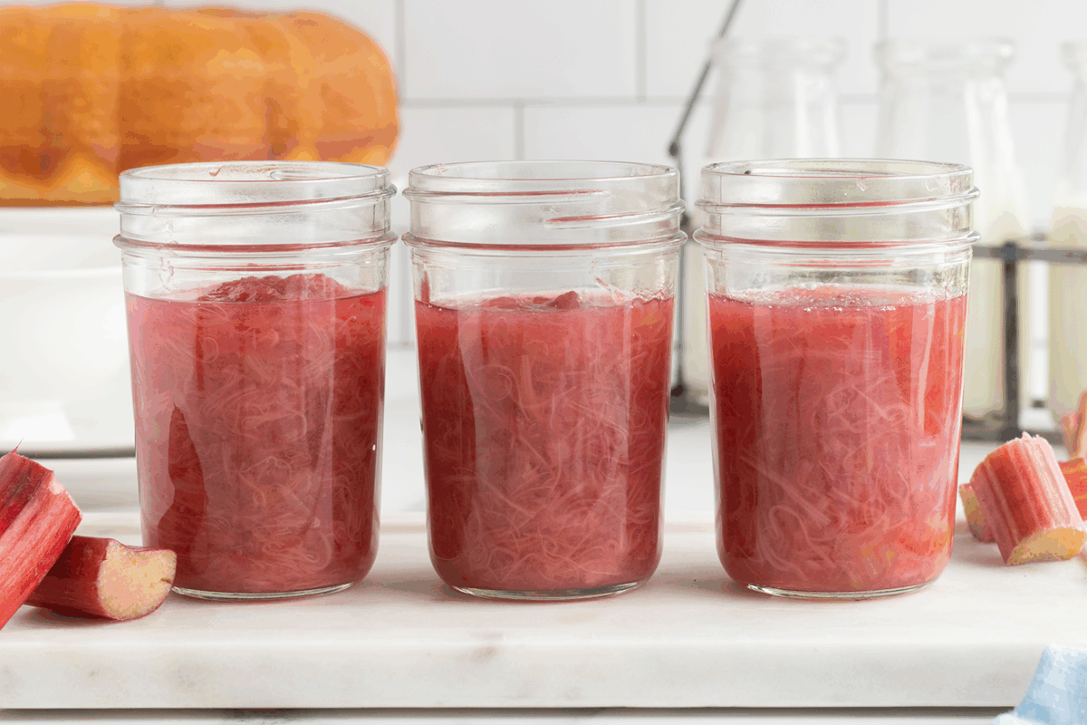 Three glass jars filled with homemade rhubarb compote sit on a white countertop. Pieces of fresh rhubarb and a cake in the background are visible in a bright kitchen setting.