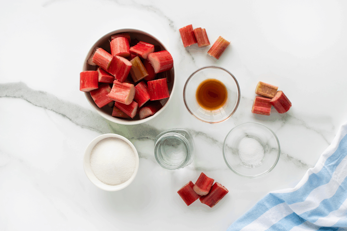 A bowl of chopped rhubarb, a small bowl of sugar, a glass of water, a dish of vanilla extract, and a small bowl of salt are arranged on a marble countertop with a blue and white striped cloth.