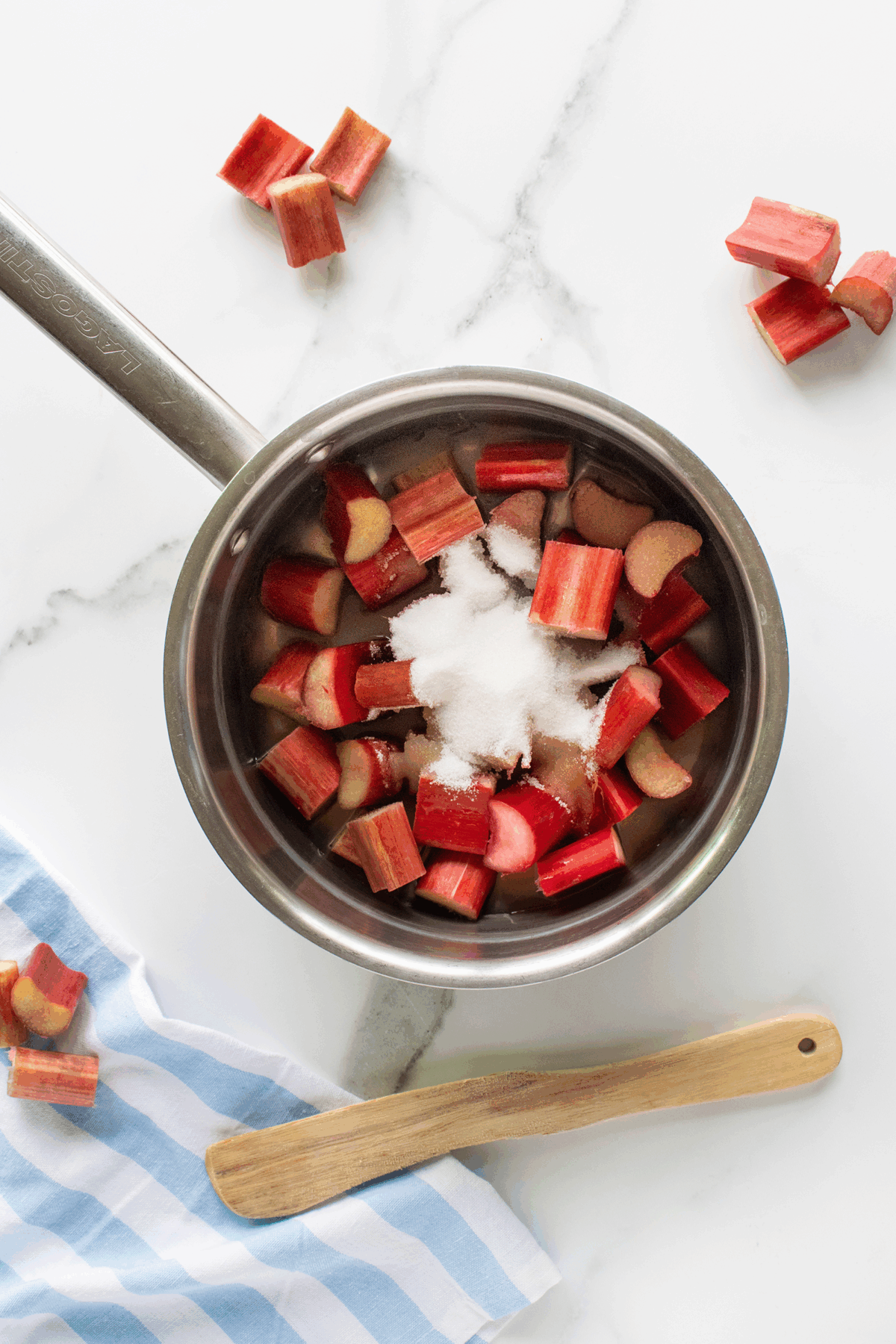 A saucepan filled with chopped rhubarb and a pile of sugar sits on a white marble surface. Nearby are extra rhubarb pieces, a wooden spatula, and a blue-striped cloth.