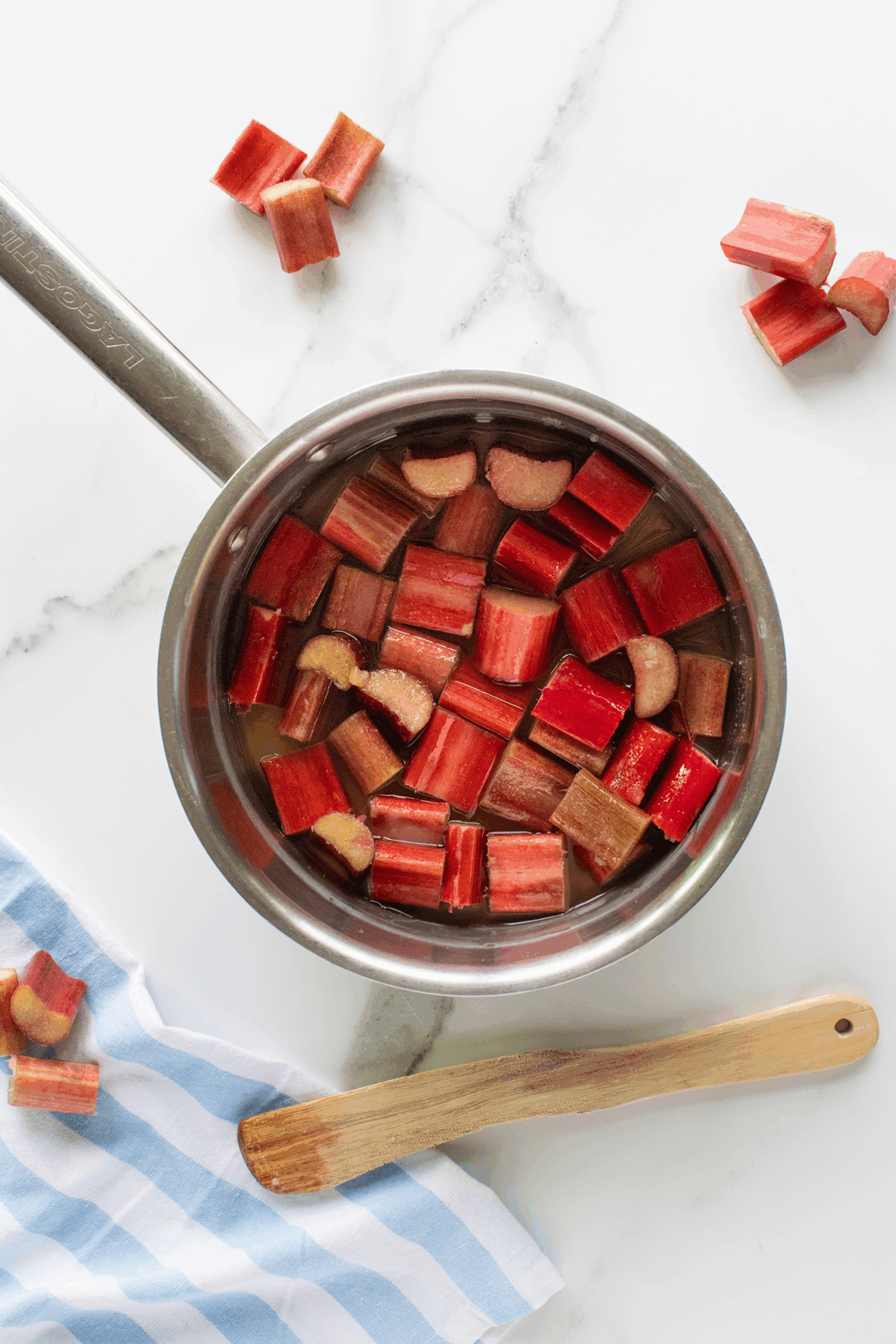 A saucepan filled with chopped rhubarb pieces and liquid sits on a white marble surface. A wooden spoon rests nearby on a blue and white striped cloth, with more rhubarb pieces scattered around.