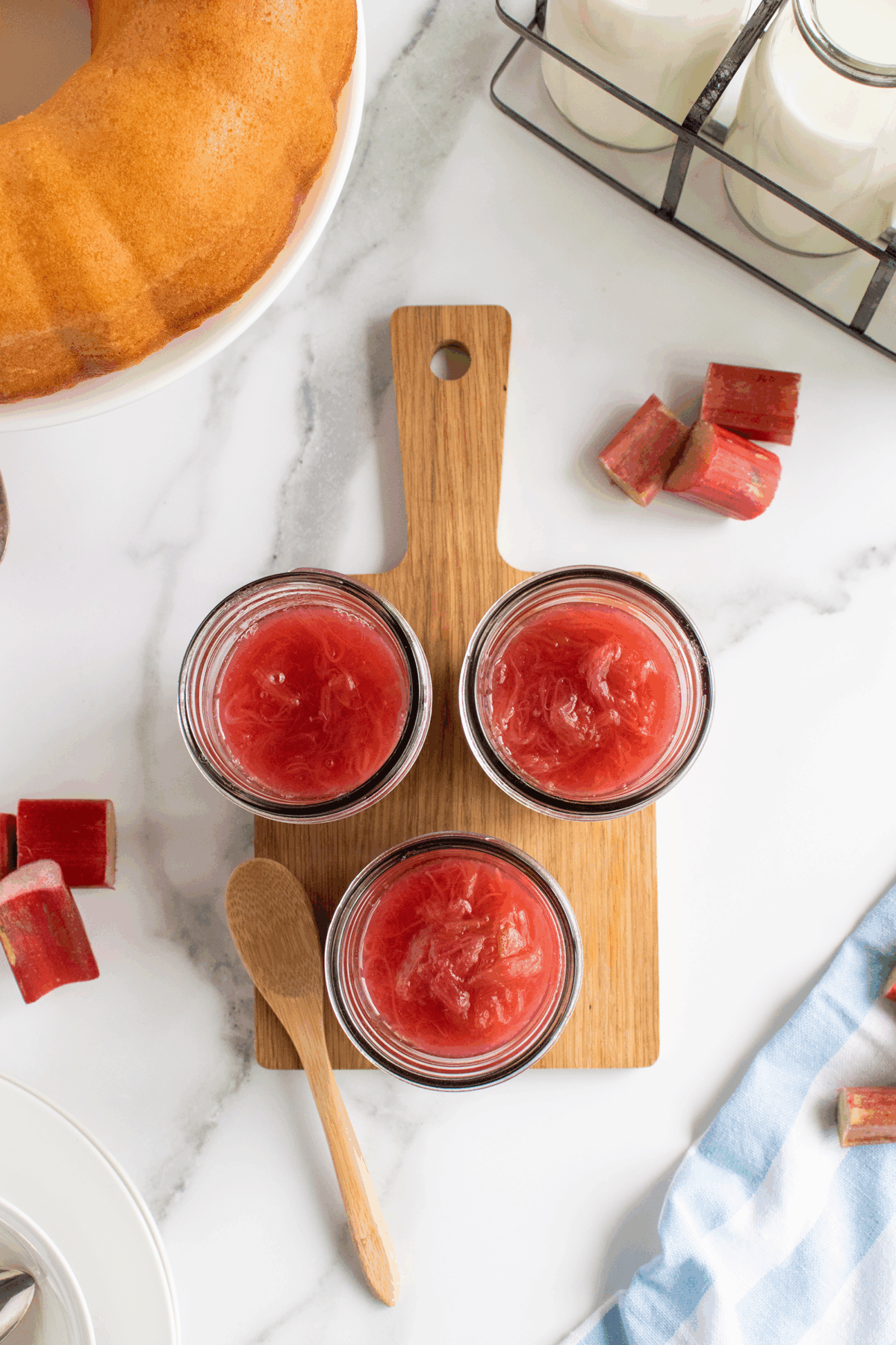 Three small glass jars filled with red rhubarb compote sit on a wooden board with a small wooden spoon. Nearby are pieces of fresh rhubarb, a Bundt cake, a blue cloth, and bottles of milk on a white surface.