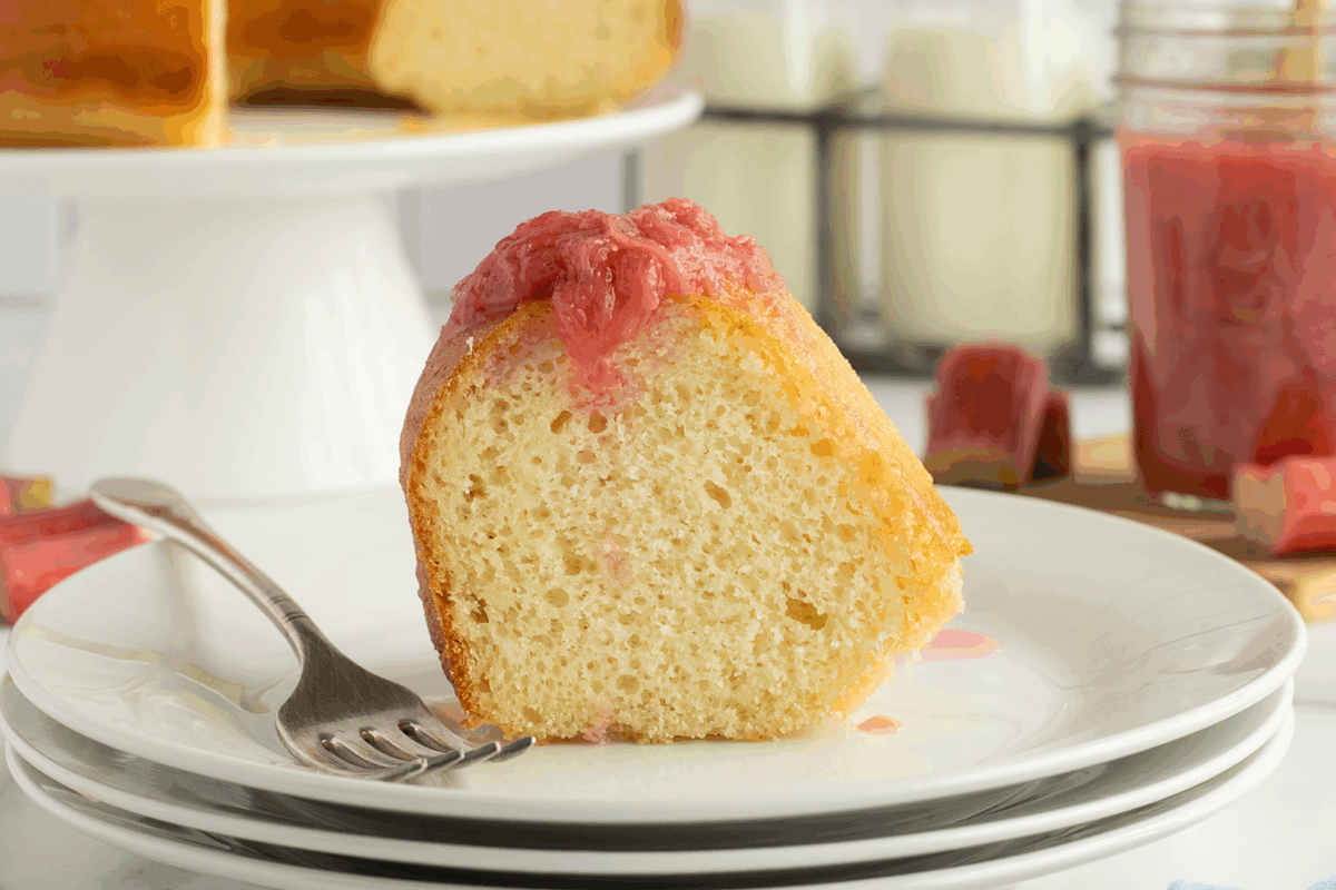 A slice of bundt cake topped with pink rhubarb sauce sits on a white plate with a fork. More cake, a jar of sauce, and glasses of milk are visible in the background.
