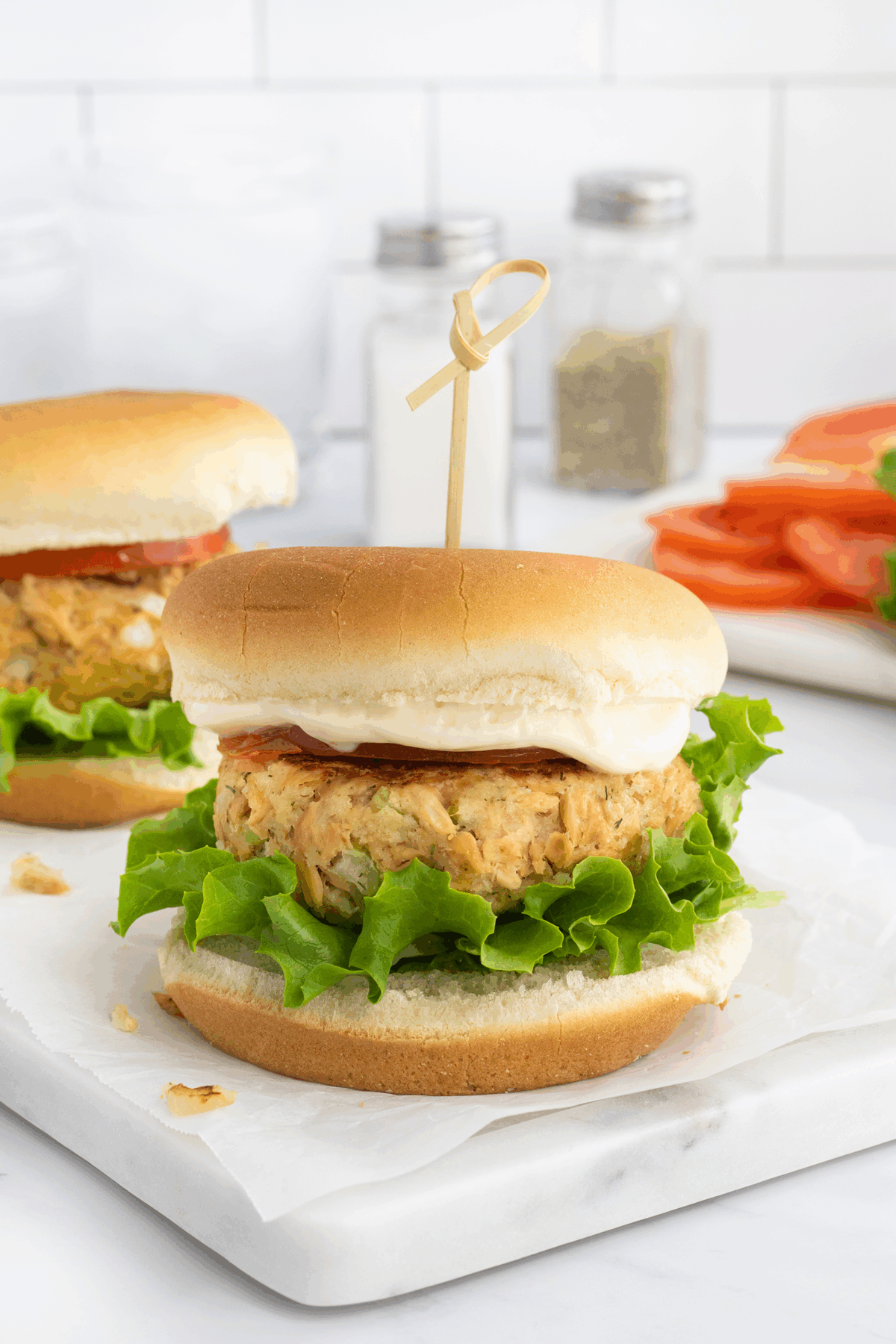 A crab cake sandwich on a bun with lettuce, tomato, and mayonnaise, secured with a wooden skewer, sits on a marble surface. Another sandwich, sliced tomatoes, and salt and pepper shakers are visible in the background.