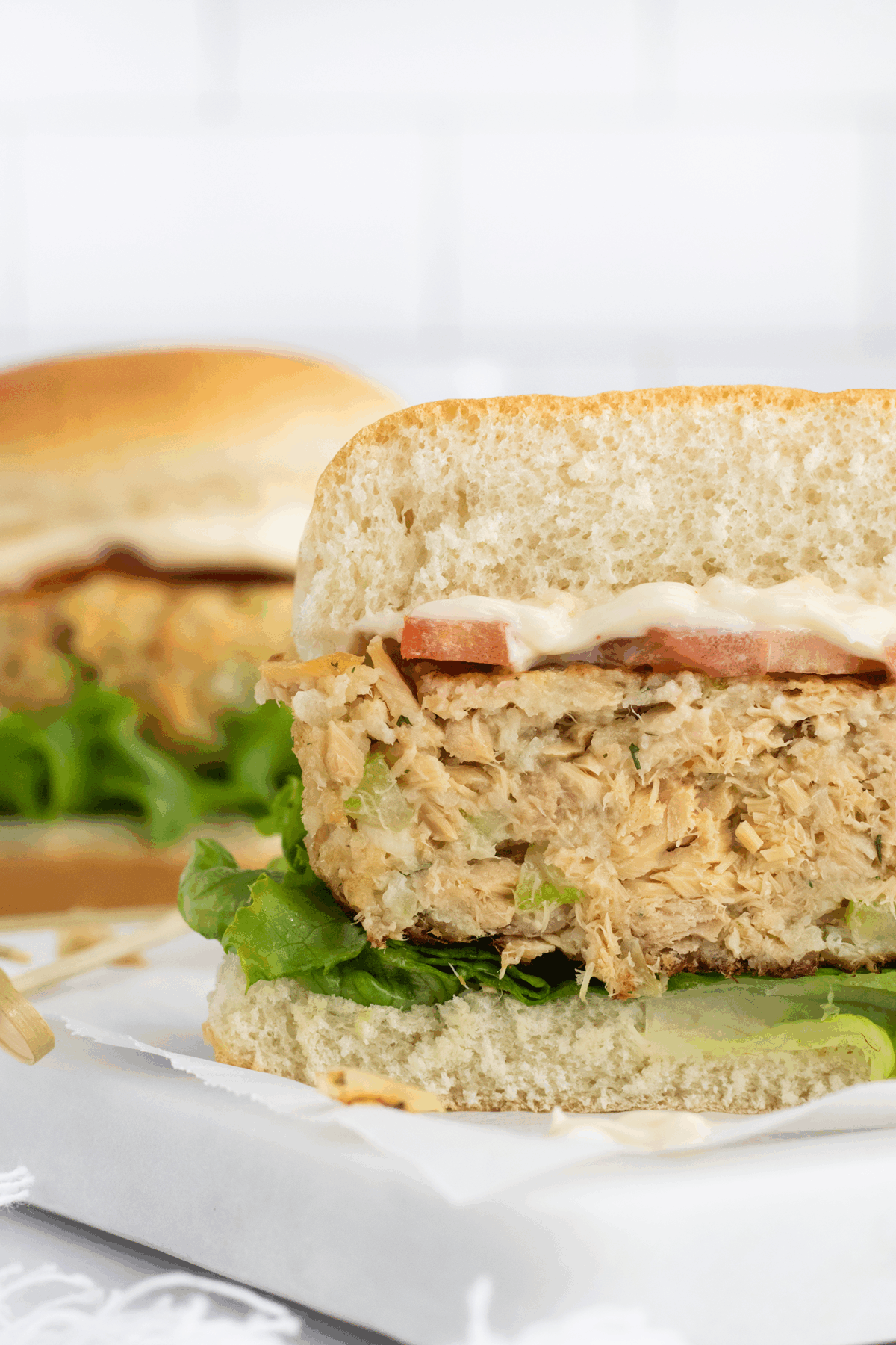 A close-up of a sandwich with a thick, textured patty, lettuce, tomato, and mayonnaise on a bun, with another similar sandwich blurred in the background.