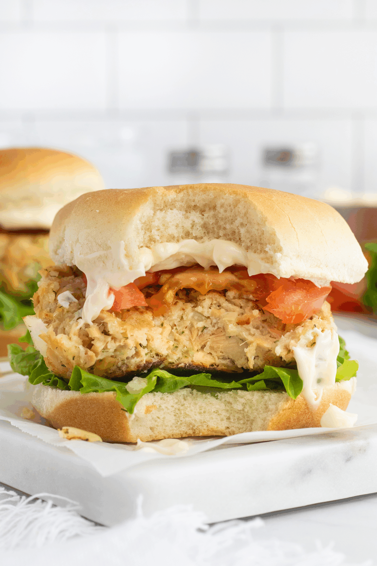 A chicken burger with lettuce, tomato, and mayonnaise on a bun, shown with a large bite taken out, sitting on a white surface with a blurred kitchen background.
