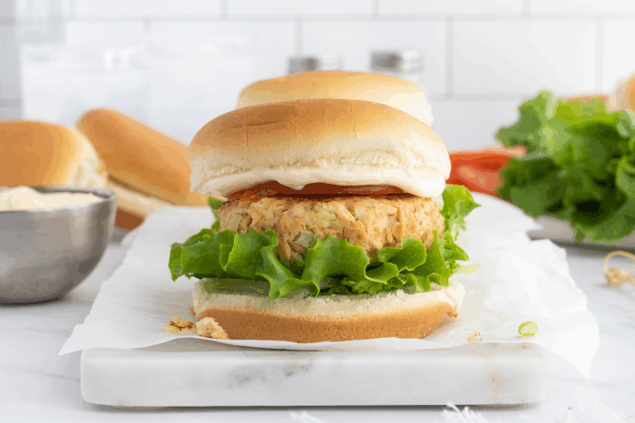 A sandwich with lettuce, a thick veggie or chicken patty, tomato, and mayonnaise in a soft bun sits on parchment paper, with lettuce, tomato, and another bun in the background.