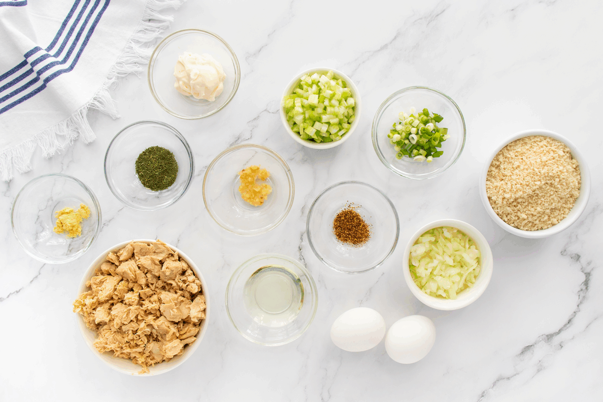 A top-down view of bowls filled with various ingredients, including chopped celery, green onions, diced onion, breadcrumbs, canned tuna, mayonnaise, spices, lemon zest, two eggs, and a small glass of oil on a marble surface.
