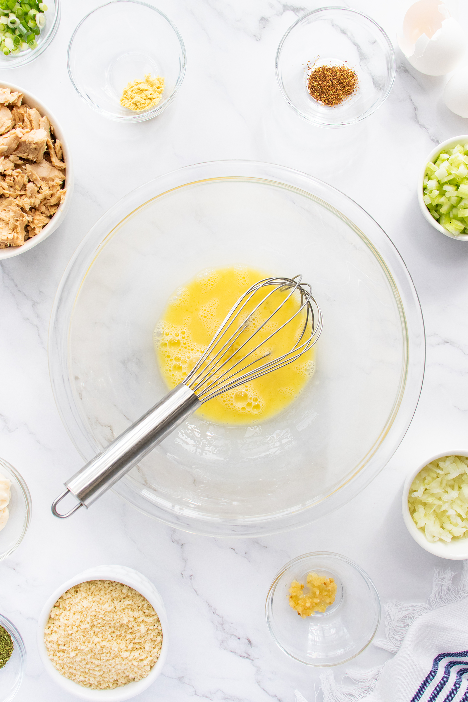A glass bowl with beaten eggs and a whisk sits on a marble surface, surrounded by small bowls of chopped vegetables, spices, and other ingredients for cooking.