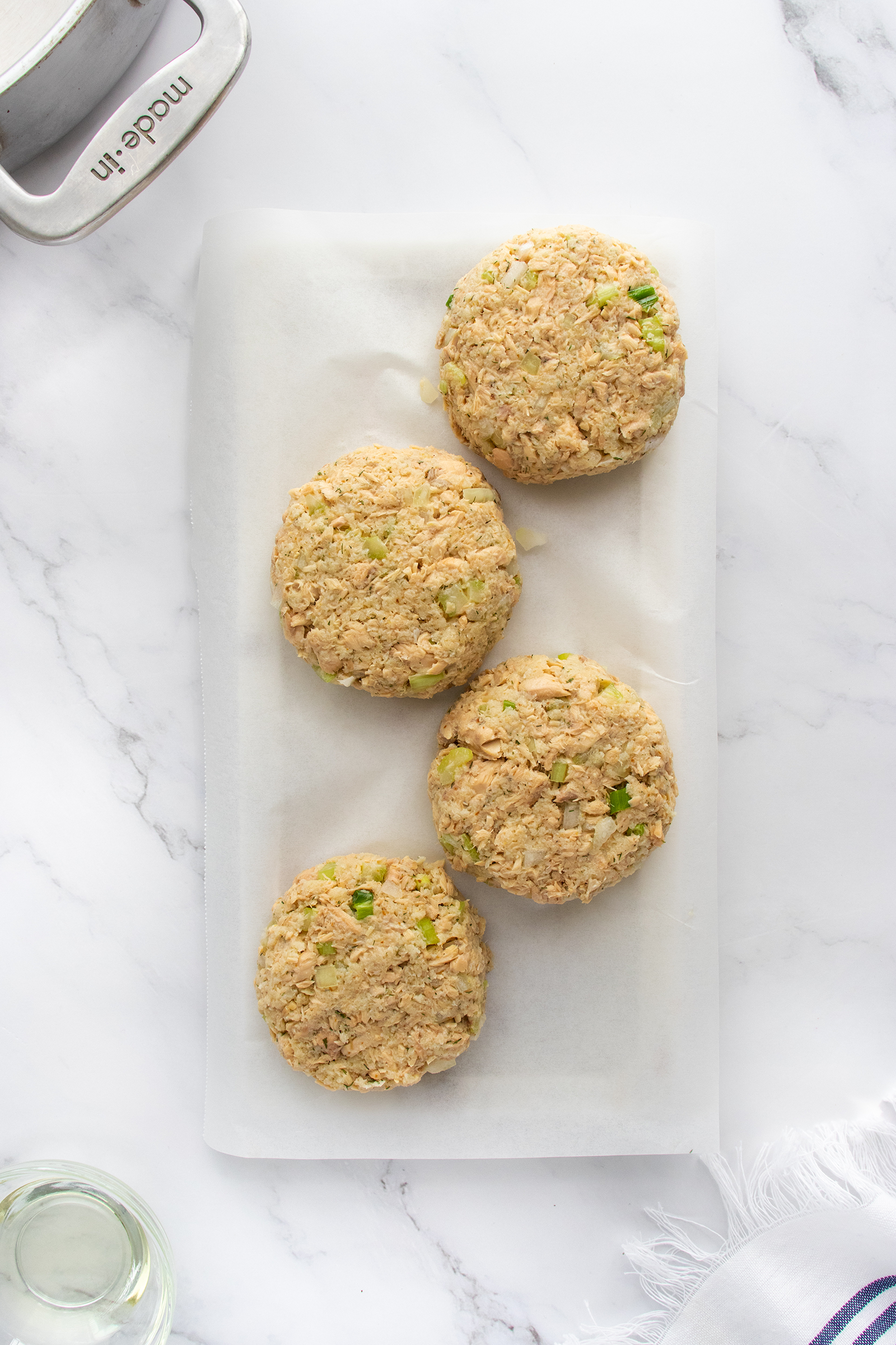 Four uncooked chickpea patties with visible green peas rest on a rectangular sheet of parchment paper on a white marble surface. A metal measuring cup and part of a glass are visible nearby.