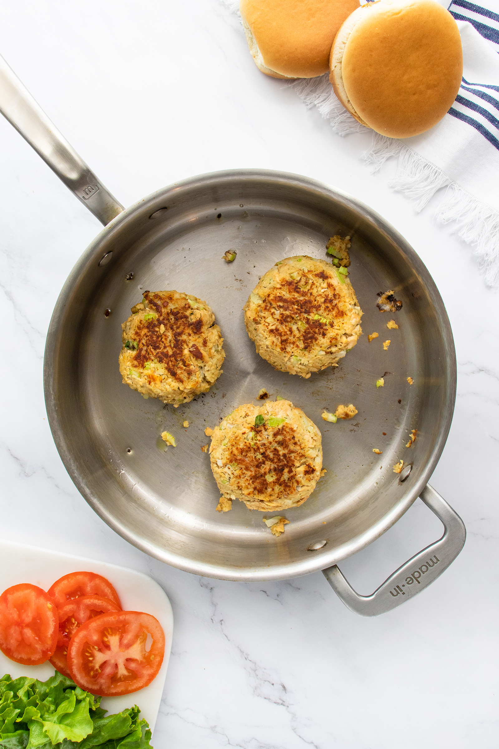 A stainless steel skillet with three golden-brown veggie burger patties. At the top, two burger buns sit on a towel. At the bottom left, sliced tomatoes and lettuce are visible on a white plate.