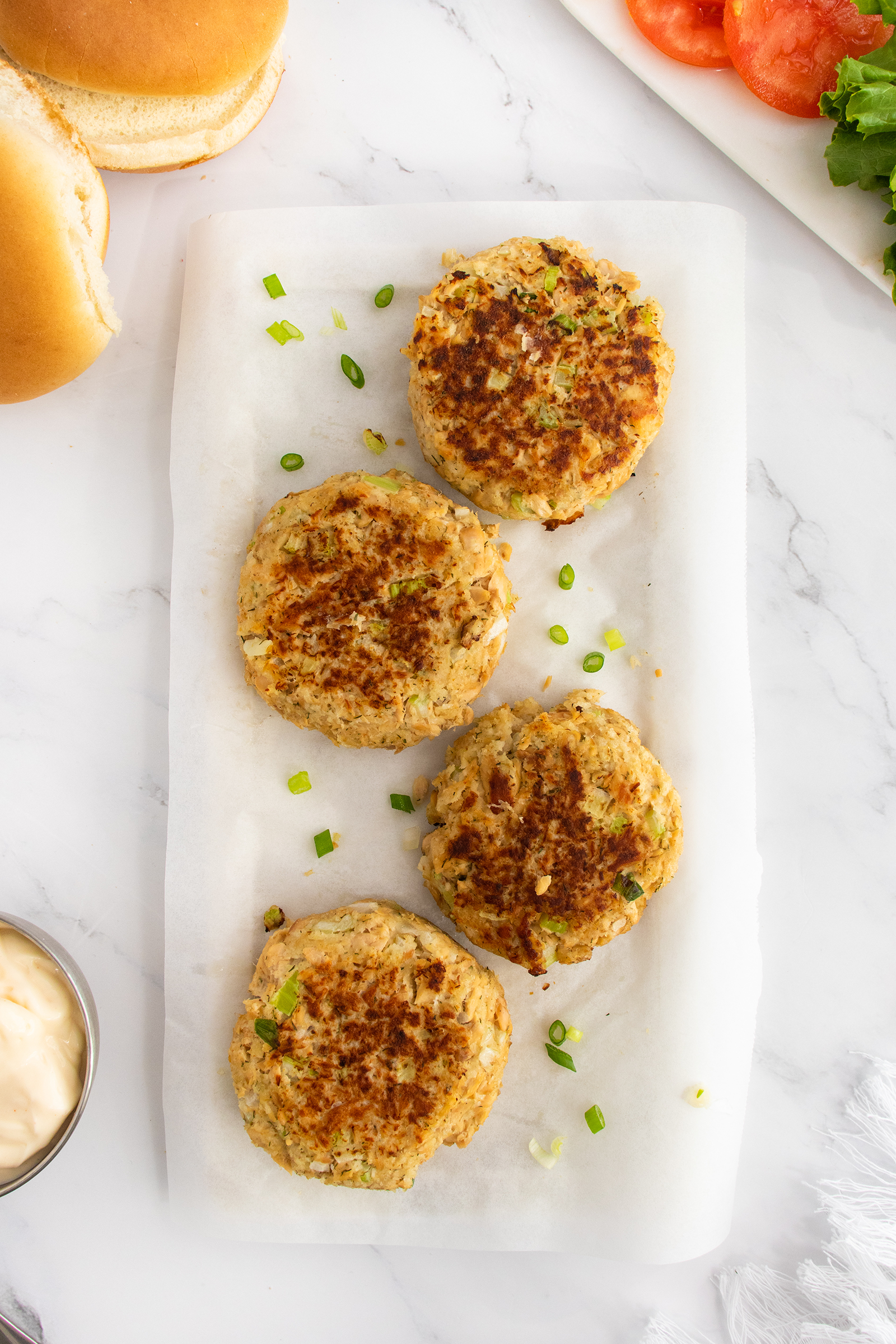 Four golden-brown crab cakes garnished with chopped green onions are arranged on white parchment paper, with sandwich buns, tomato slices, lettuce, and a bowl of mayonnaise nearby on a marble surface.