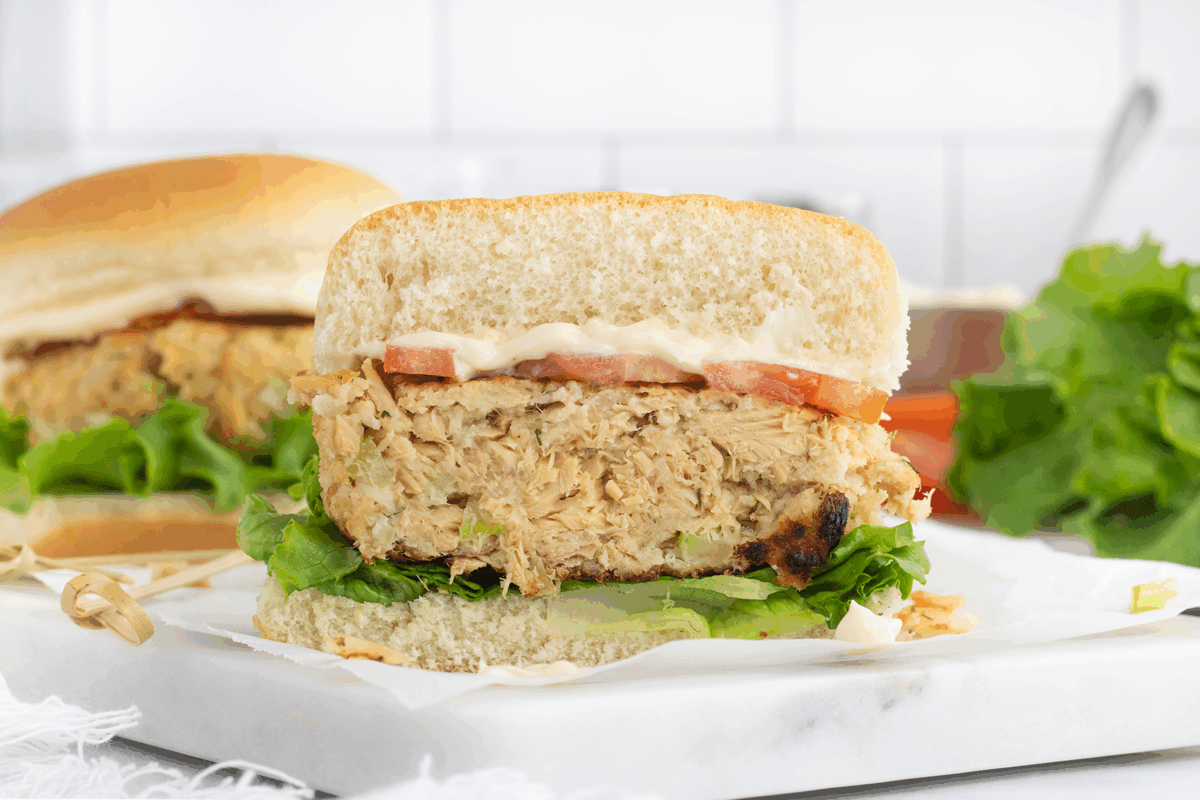 A sandwich with a thick, flaky patty, leafy green lettuce, tomato slices, and mayonnaise in a soft bun is shown cut in half. Another sandwich and loose lettuce are visible in the background.
