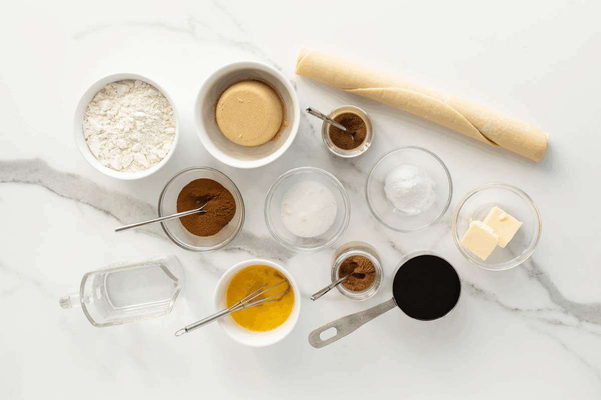 Various baking ingredients arranged on a white marble surface, including flour, brown sugar, butter, spices, egg wash, rolled-out dough, vanilla extract, molasses, and a cup of syrup, all in small bowls or measuring cups.