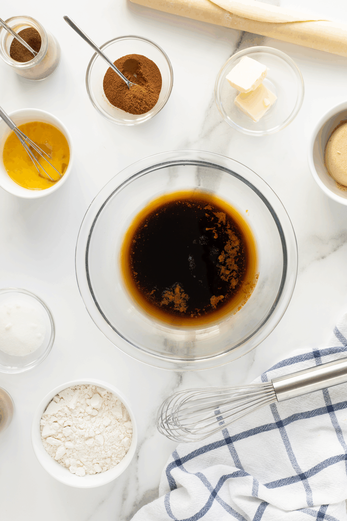 A glass bowl with dark liquid surrounded by small bowls containing flour, sugar, brown sugar, butter, cinnamon, egg mixture, and other baking ingredients on a white surface with a whisk and kitchen towel nearby.