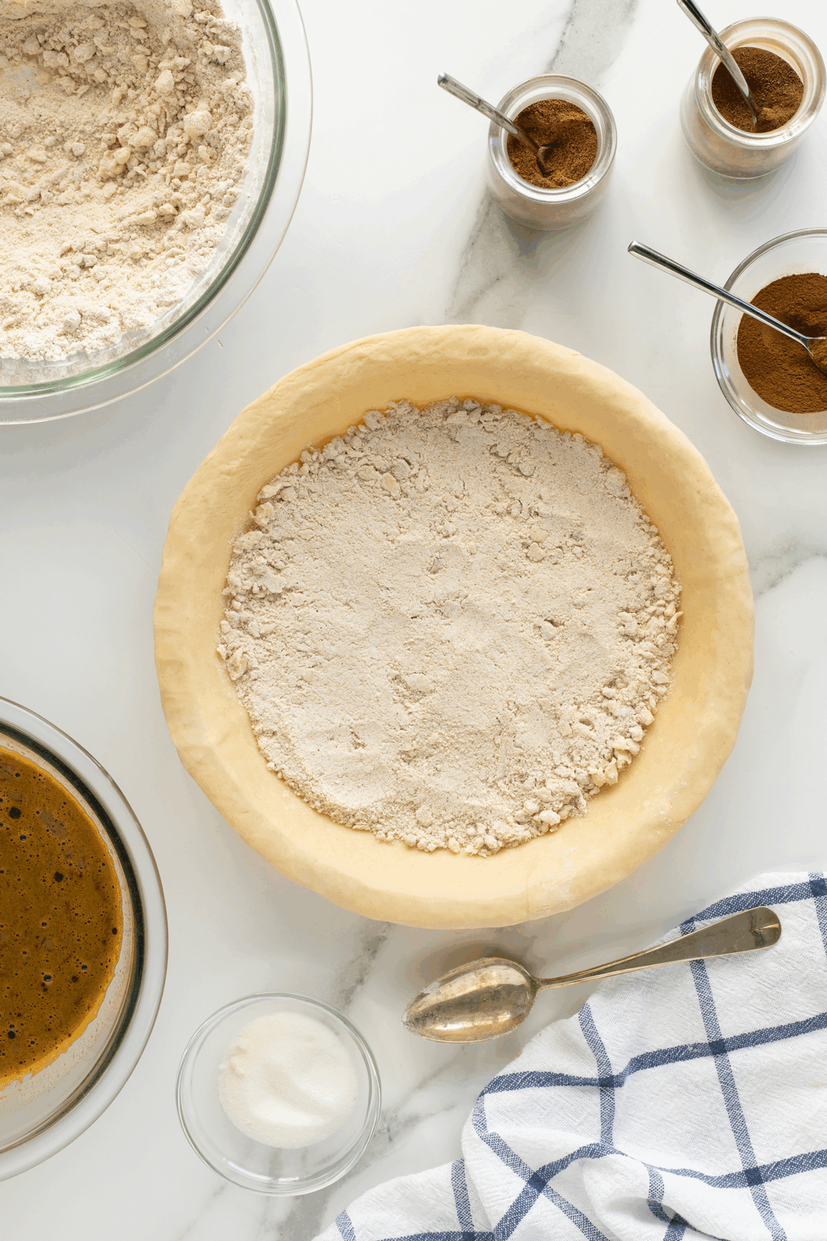 A pie crust filled with a layer of flour mixture sits on a white surface, surrounded by bowls of ingredients, small jars of spices with spoons, a striped kitchen towel, and a spoon.