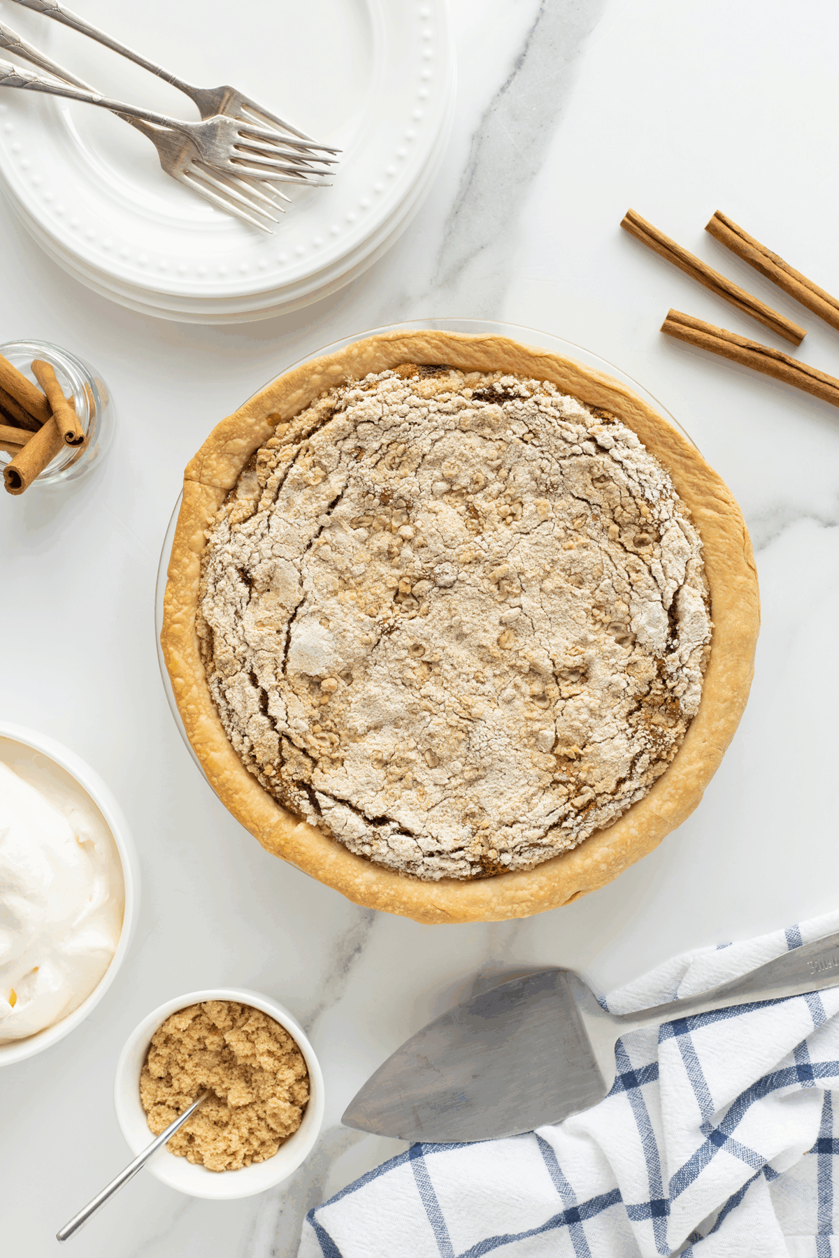 A freshly baked pie with a cracked, golden-brown crust sits on a marble surface surrounded by cinnamon sticks, a jar of cinnamon, a bowl of whipped cream, brown sugar, plates, forks, and a striped kitchen towel.
