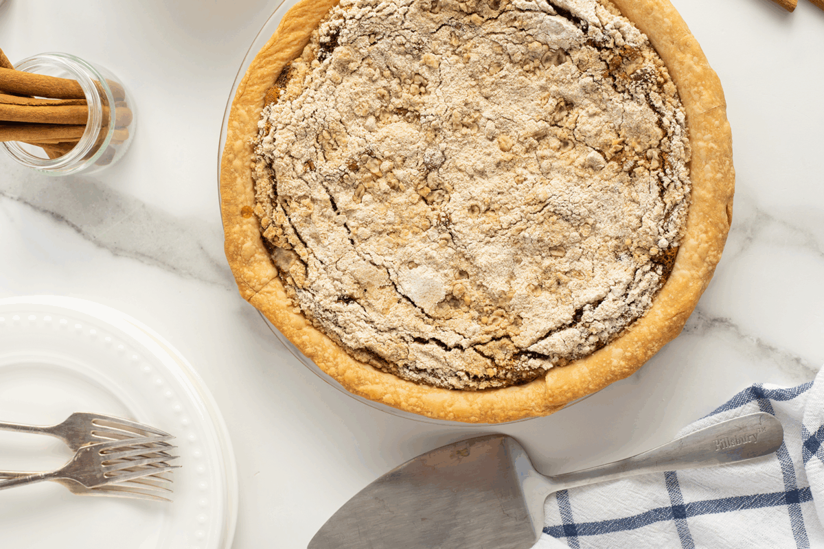 A baked pie with a crumbly topping sits on a marble surface next to a pie server, a blue-and-white cloth, two forks on a white plate, and a jar with cinnamon sticks.